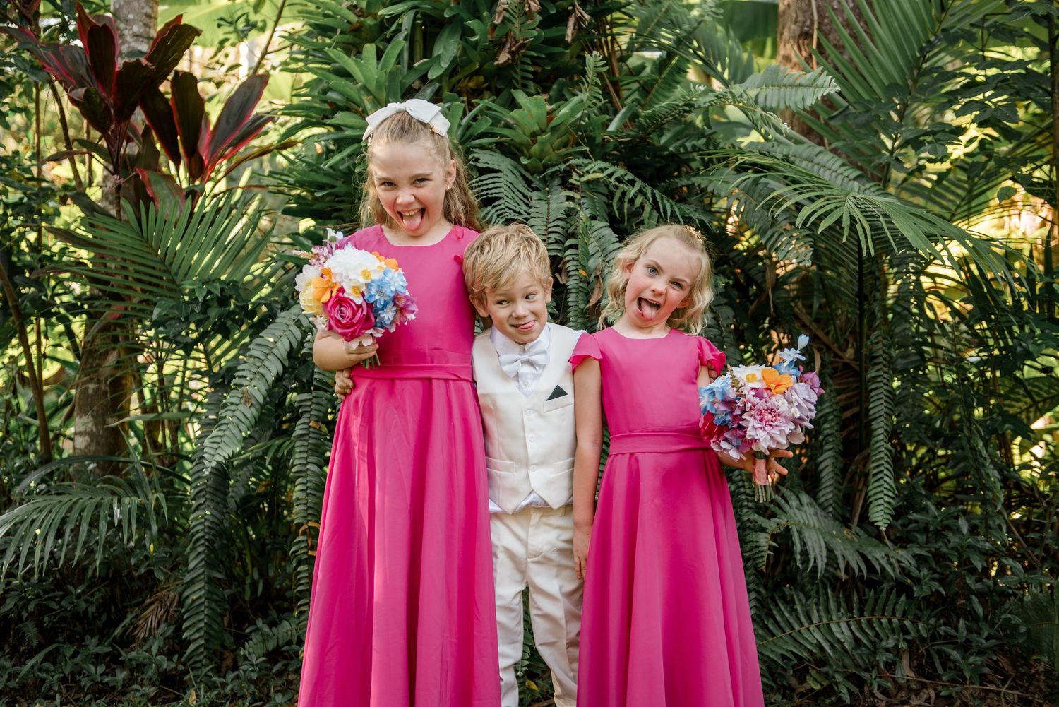 Two flower girls in pink dresses hold colorful bouquets with a ring bearer in white suit among tropical greenery.
