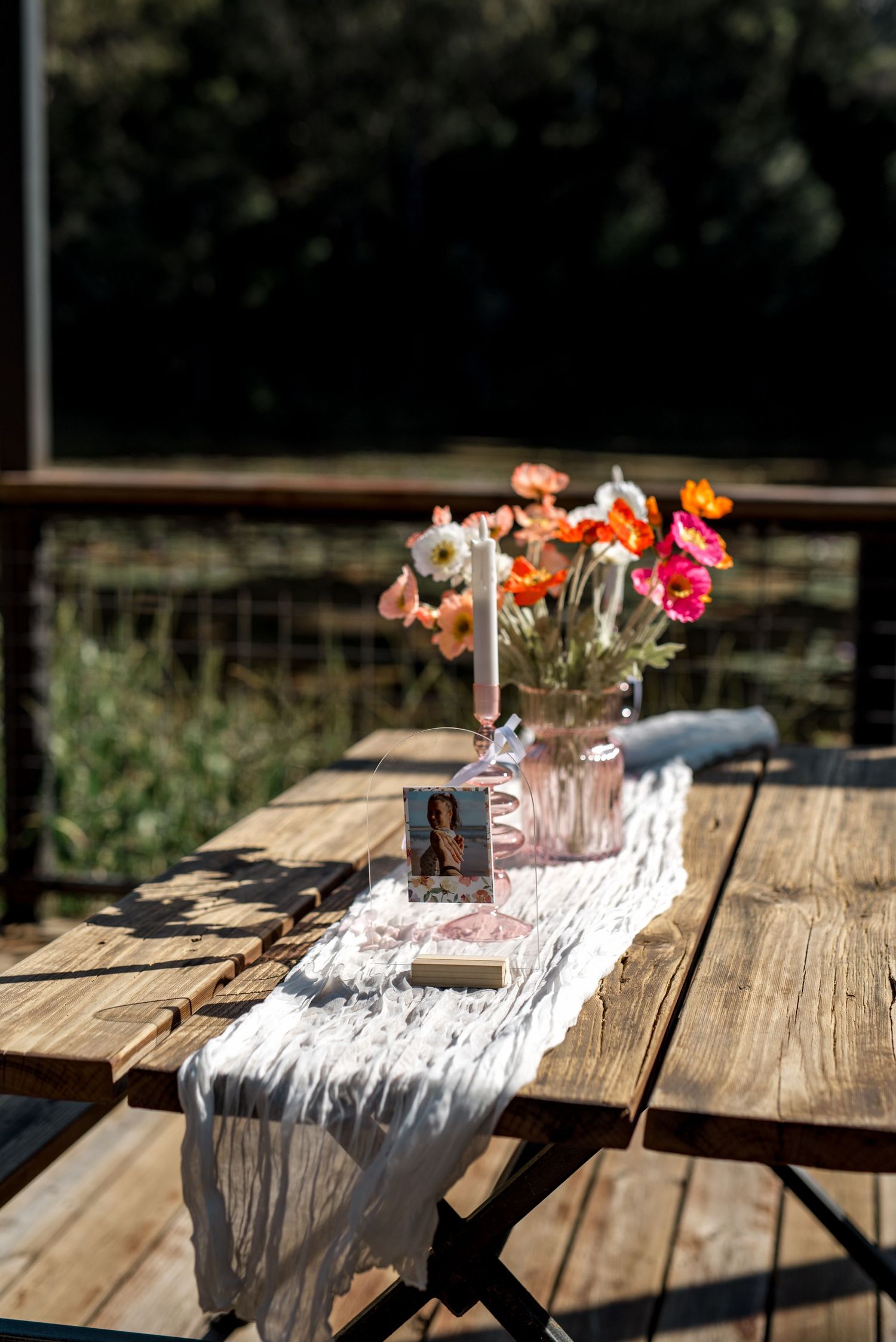Rustic wooden picnic table decorated with a white lace runner, pink candle, and bright orange flowers in a vase outdoors.