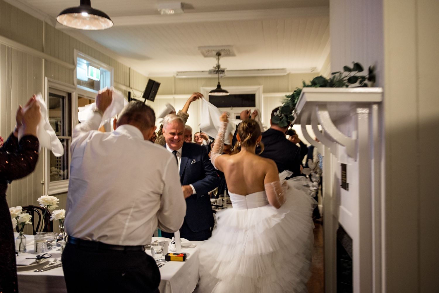 Bride in white gown dancing with guests waving napkins at a joyful wedding reception.