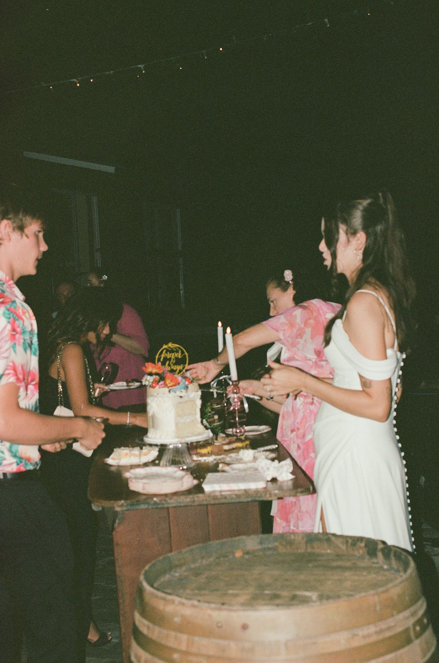 Bride in white dress cutting wedding cake at rustic barrel table with guests.