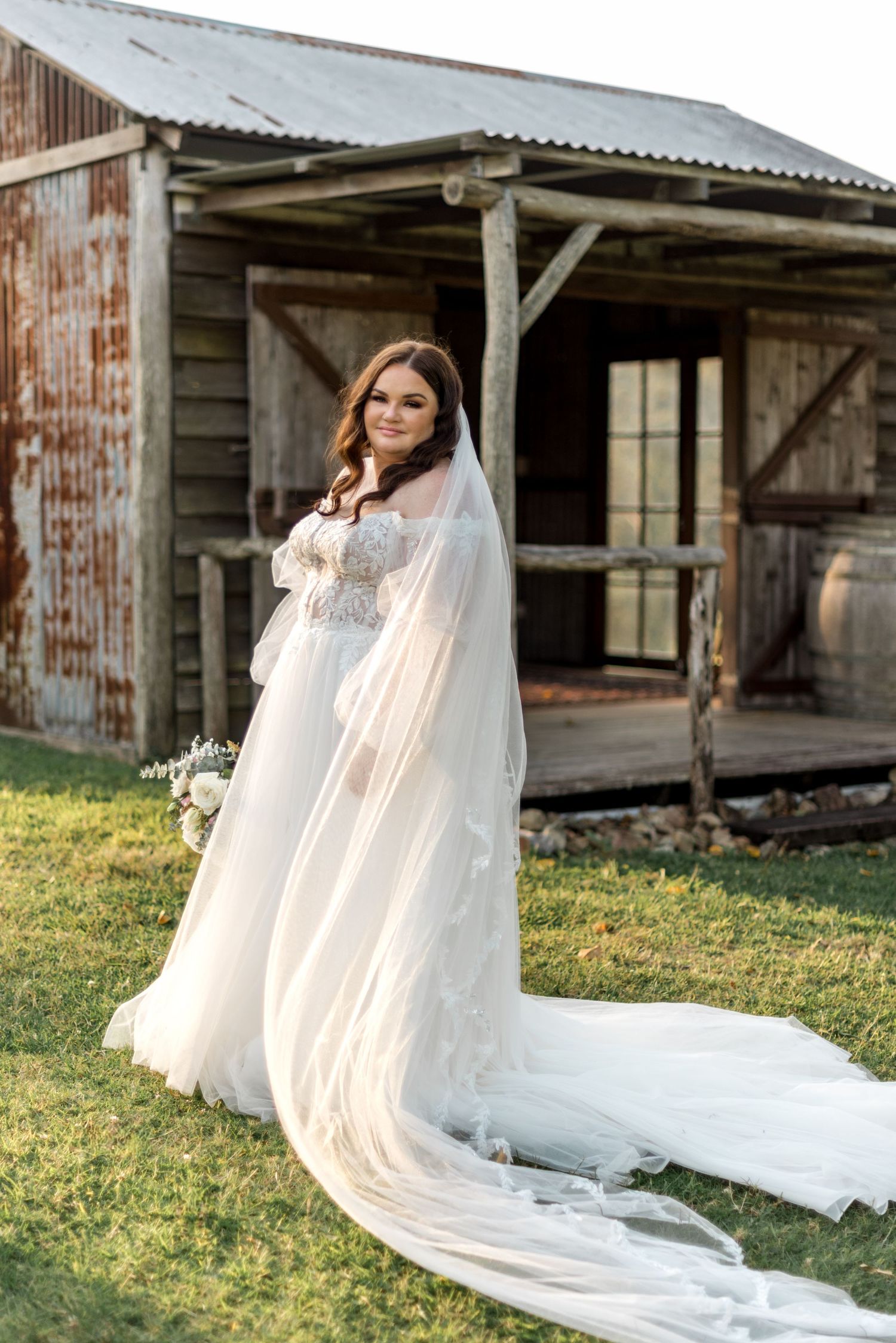 Bride in lace wedding gown with long veil posing before rustic barn at sunset.