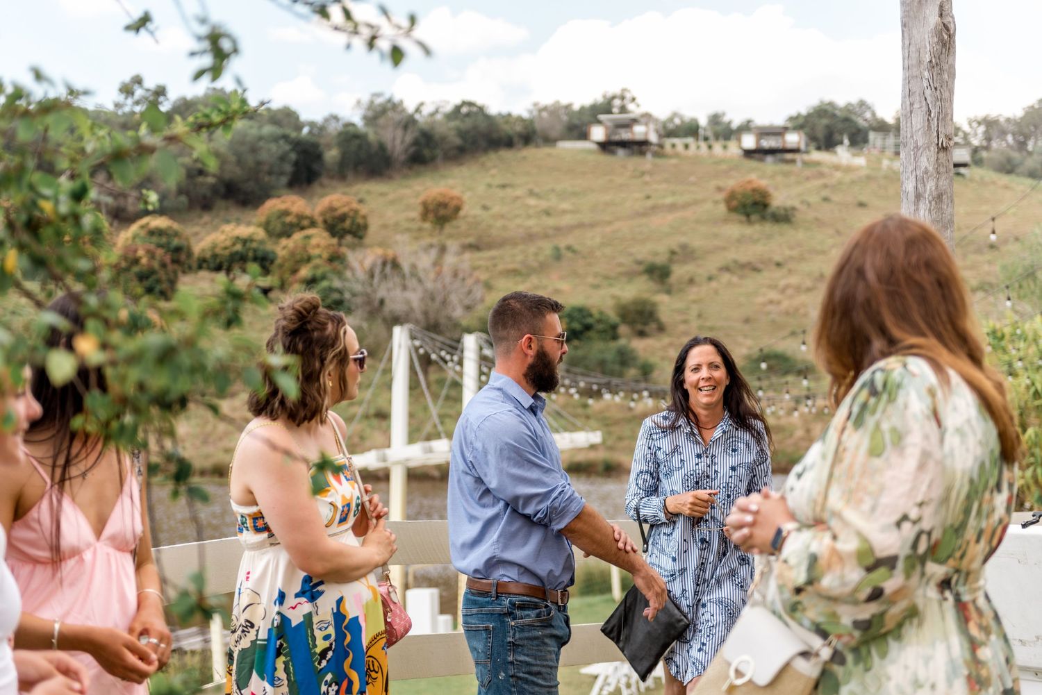 Wedding guests mingling outdoors with rolling green hills in the background at a rustic countryside celebration.