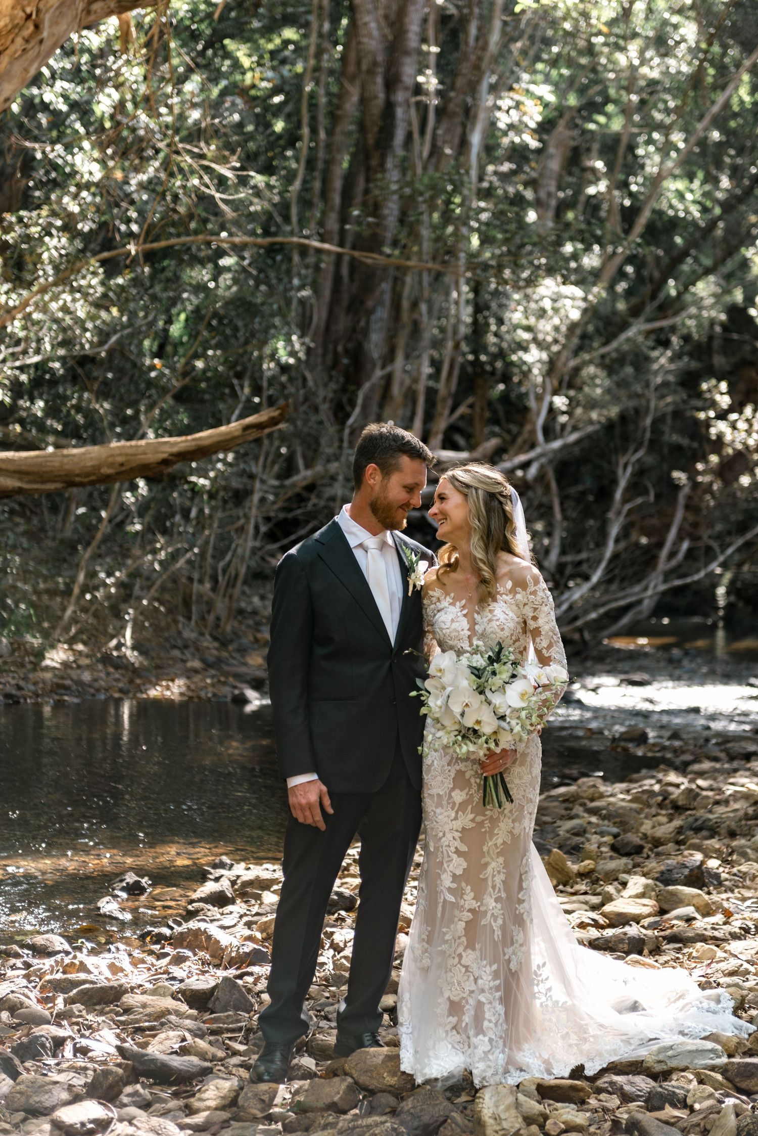 Bride and groom share romantic moment by rocky creek surrounded by lush forest.