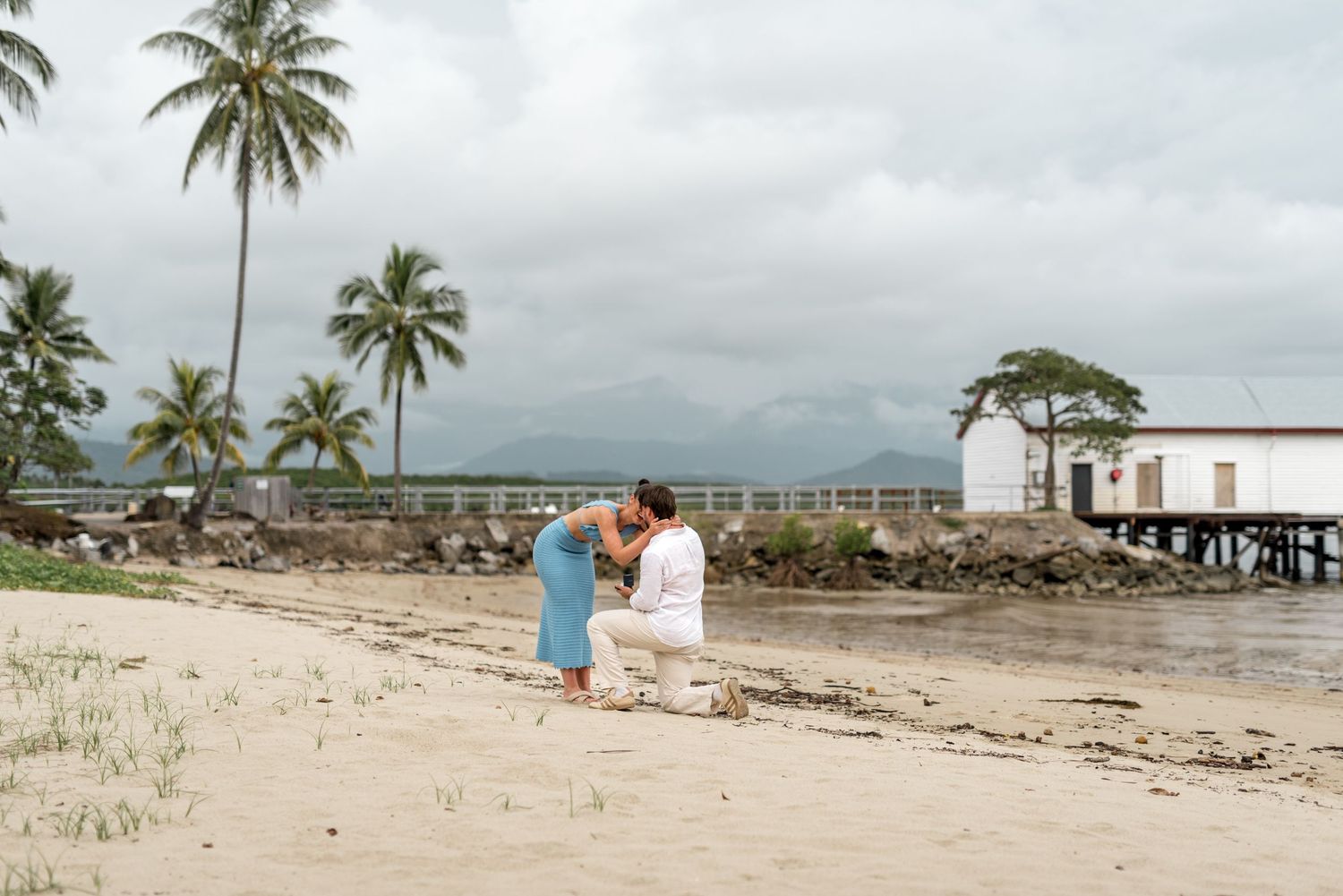 Romantic beach proposal with couple embracing on sandy shore beside tropical waterfront pier.