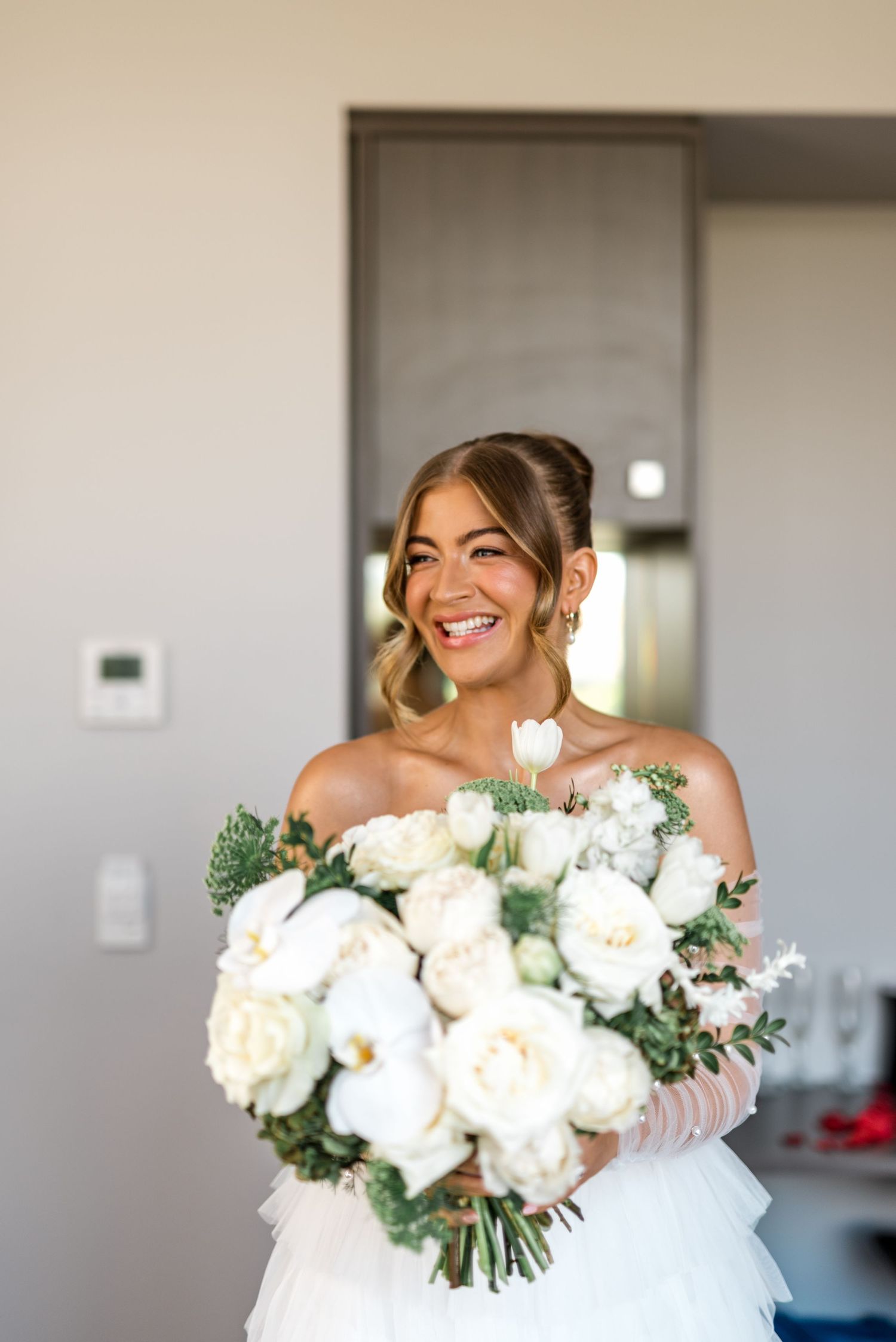 Radiant bride in ruffled wedding gown holding large white peony and rose bouquet, smiling joyfully.