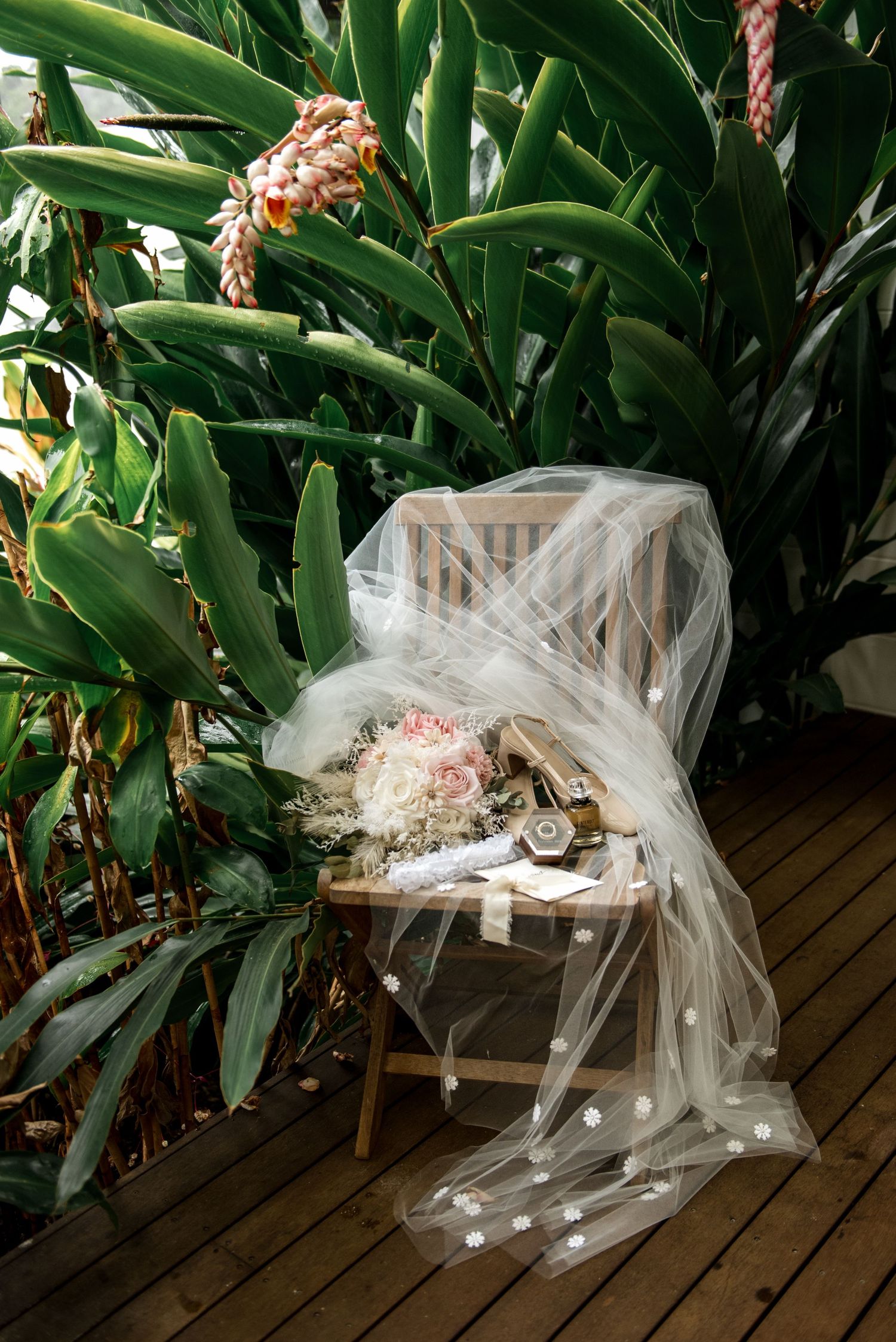 Bridal veil draped over wooden chair with bouquet and accessories surrounded by lush tropical plants.