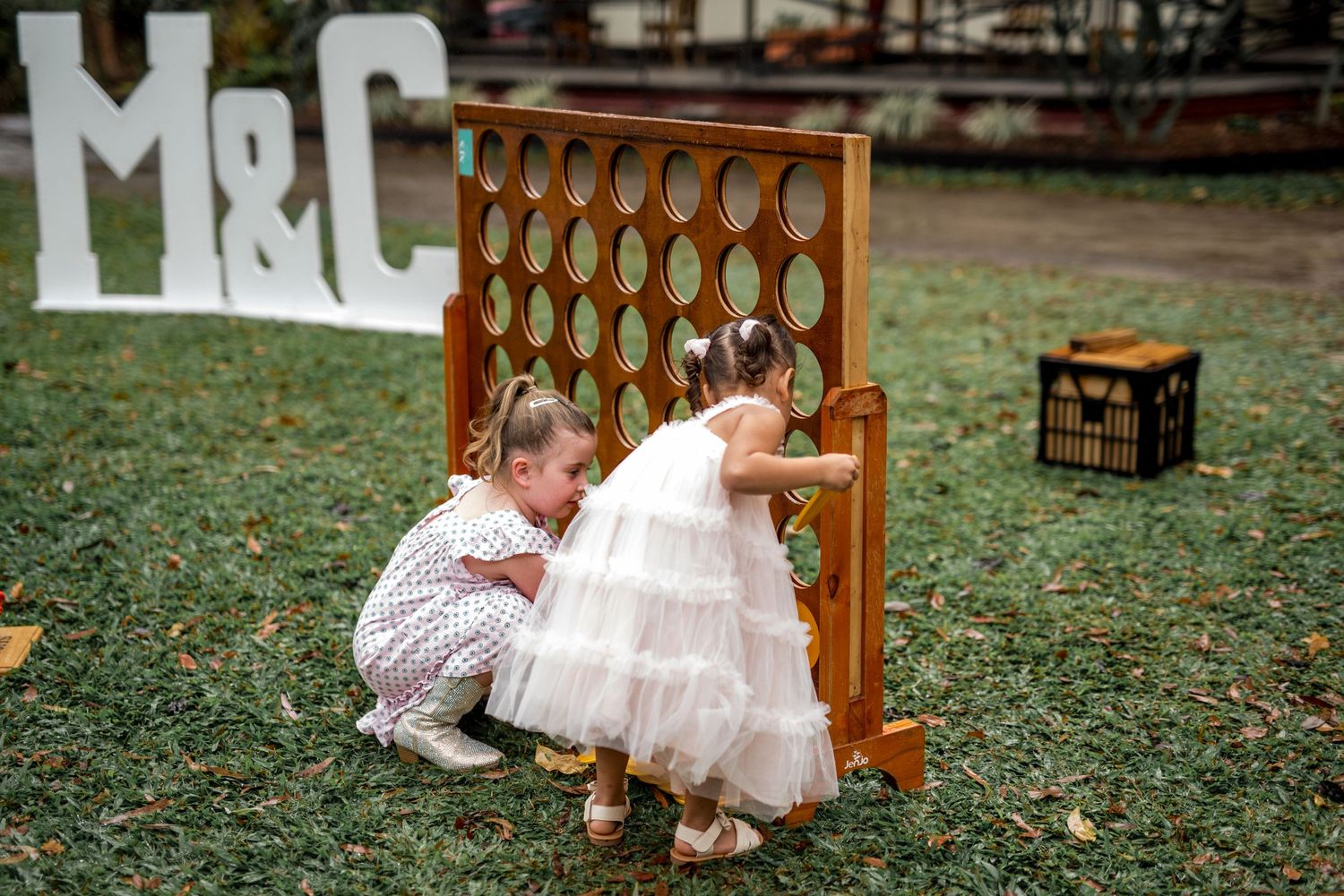 Two young flower girls playing a giant Connect Four game at an outdoor wedding reception.