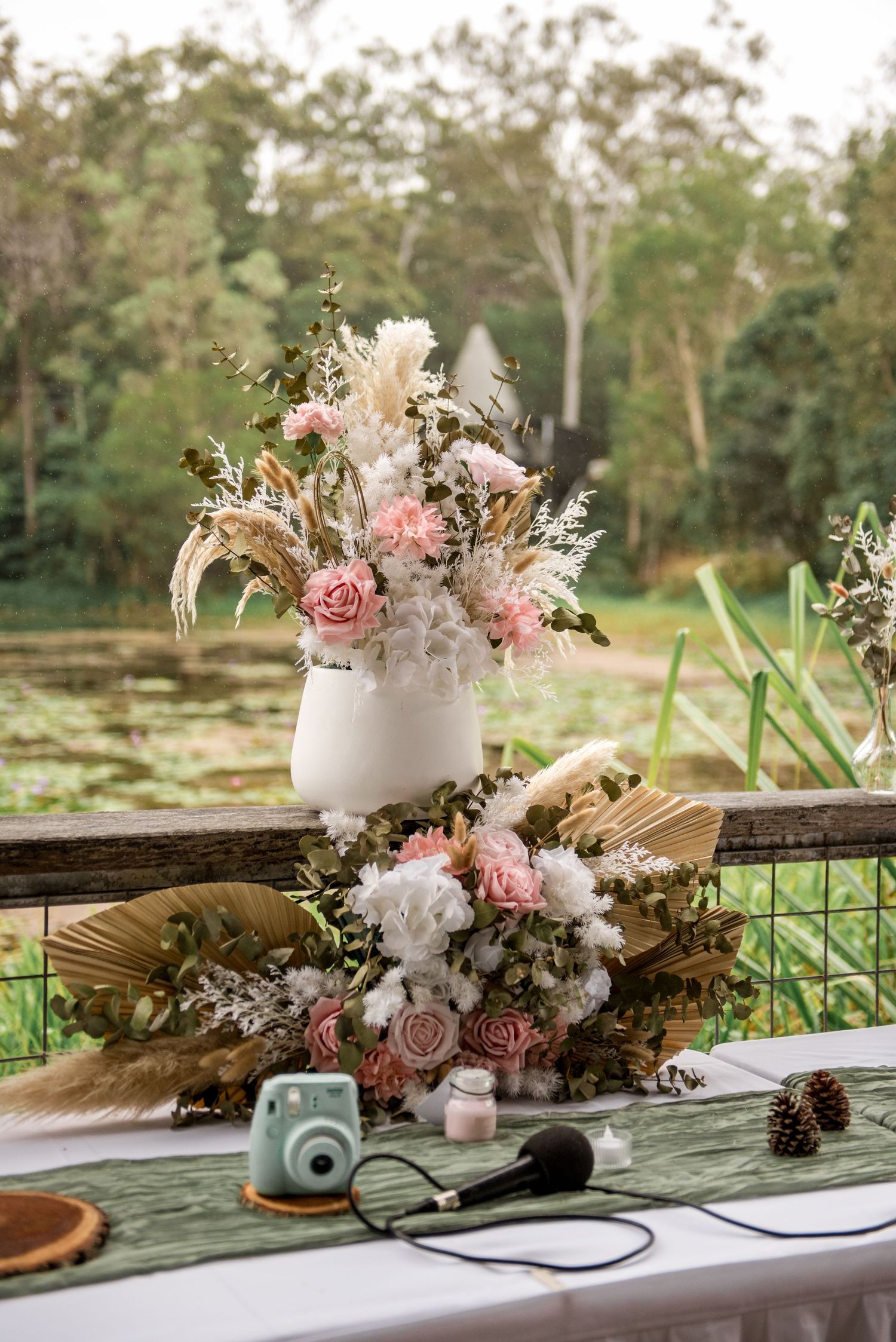 Boho wedding table centerpiece with pink roses, dried pampas grass, and white blooms near a pond.
