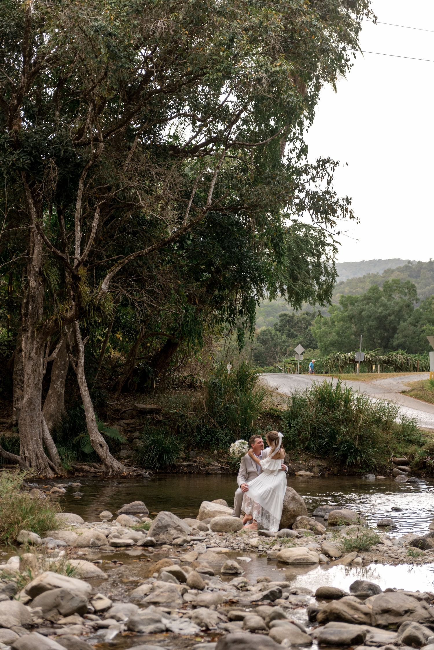 Bride and groom sit together on rocks beside a peaceful flowing creek in a natural setting.