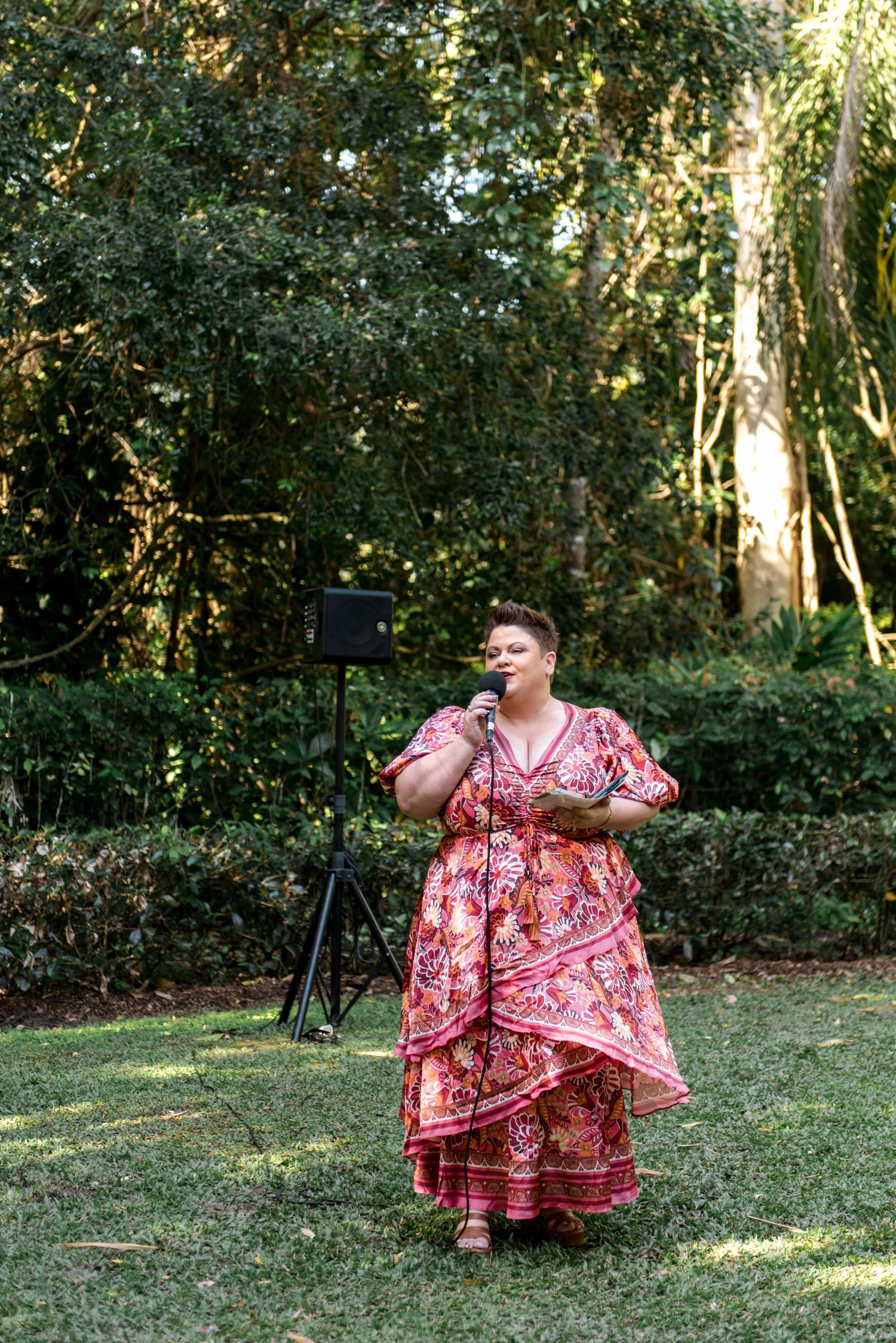 Singer in floral red dress performing with microphone at outdoor wedding reception in lush garden setting.