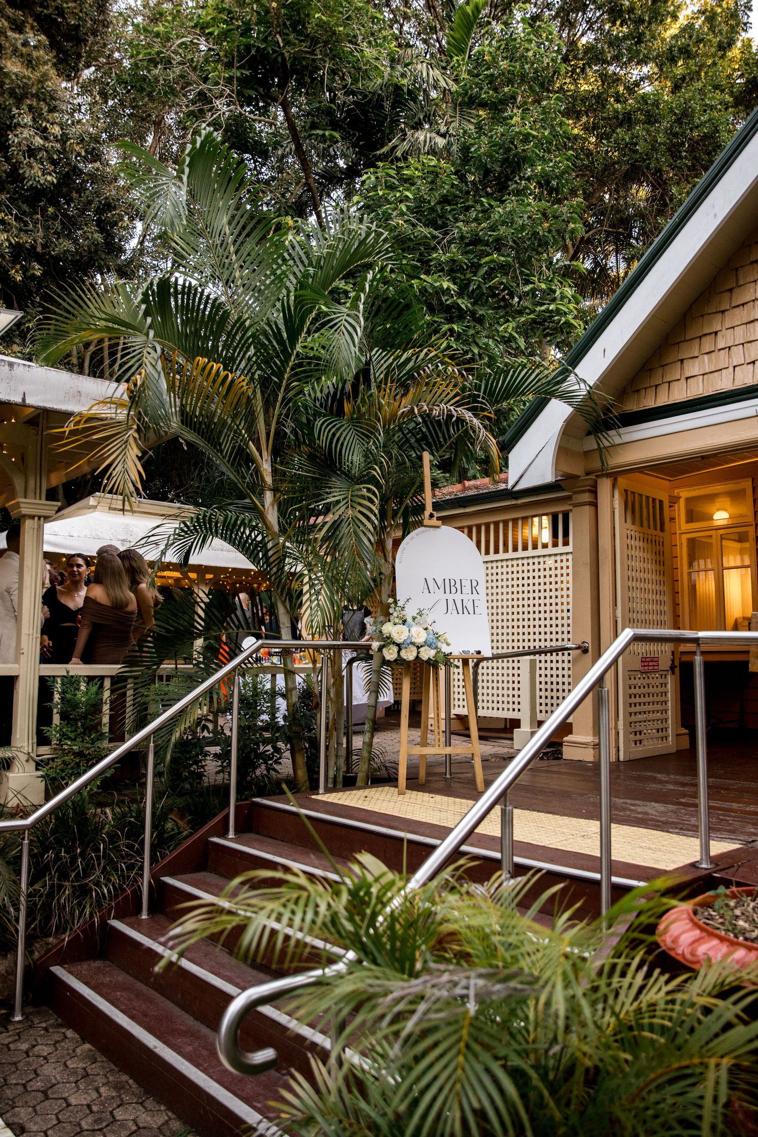 Rustic outdoor wedding venue entrance with tropical palms, fairy lights, and a welcome sign reading Amber and Jake.