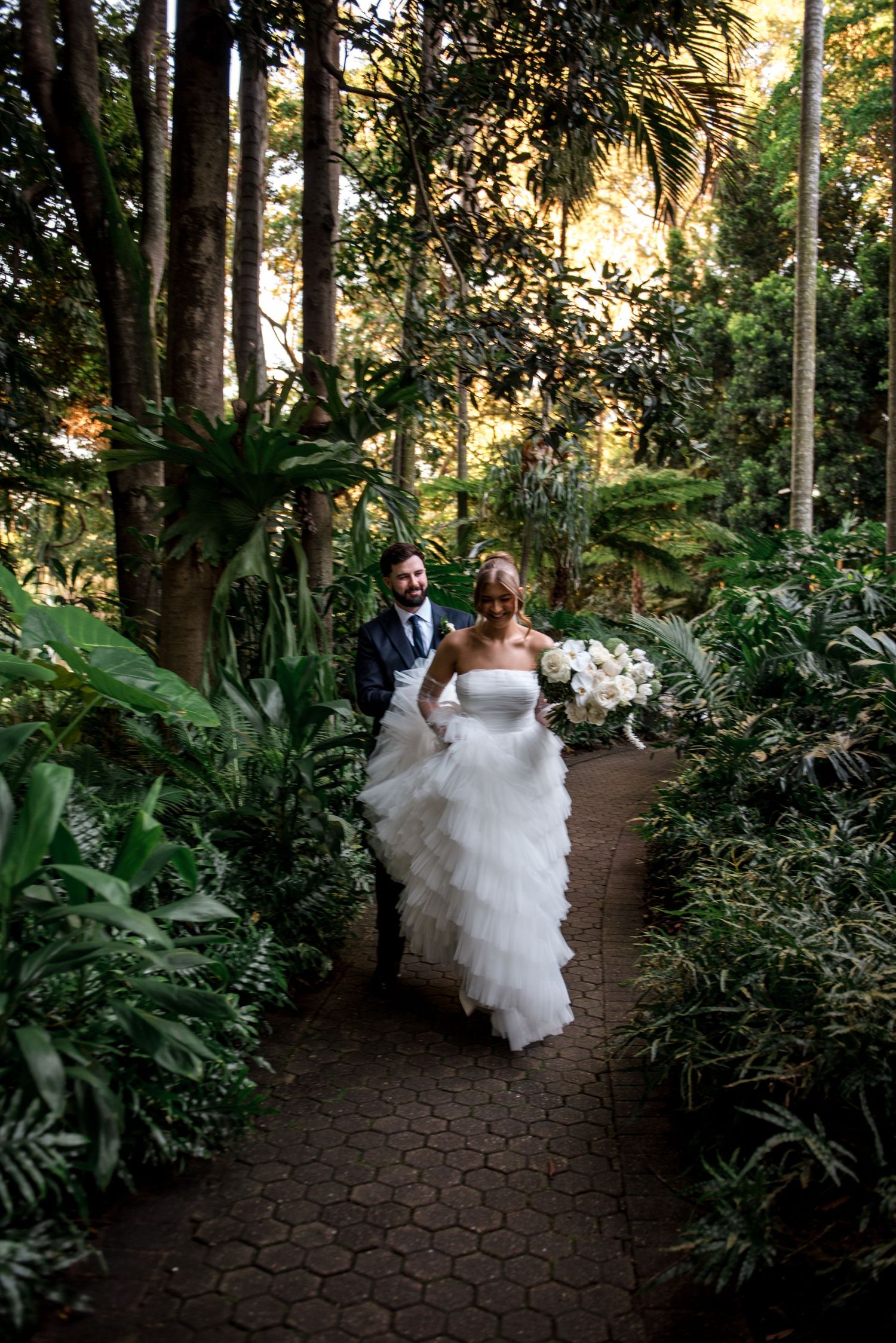Bride and groom walk hand in hand along lush garden path, bride in tiered white gown holding bouquet.