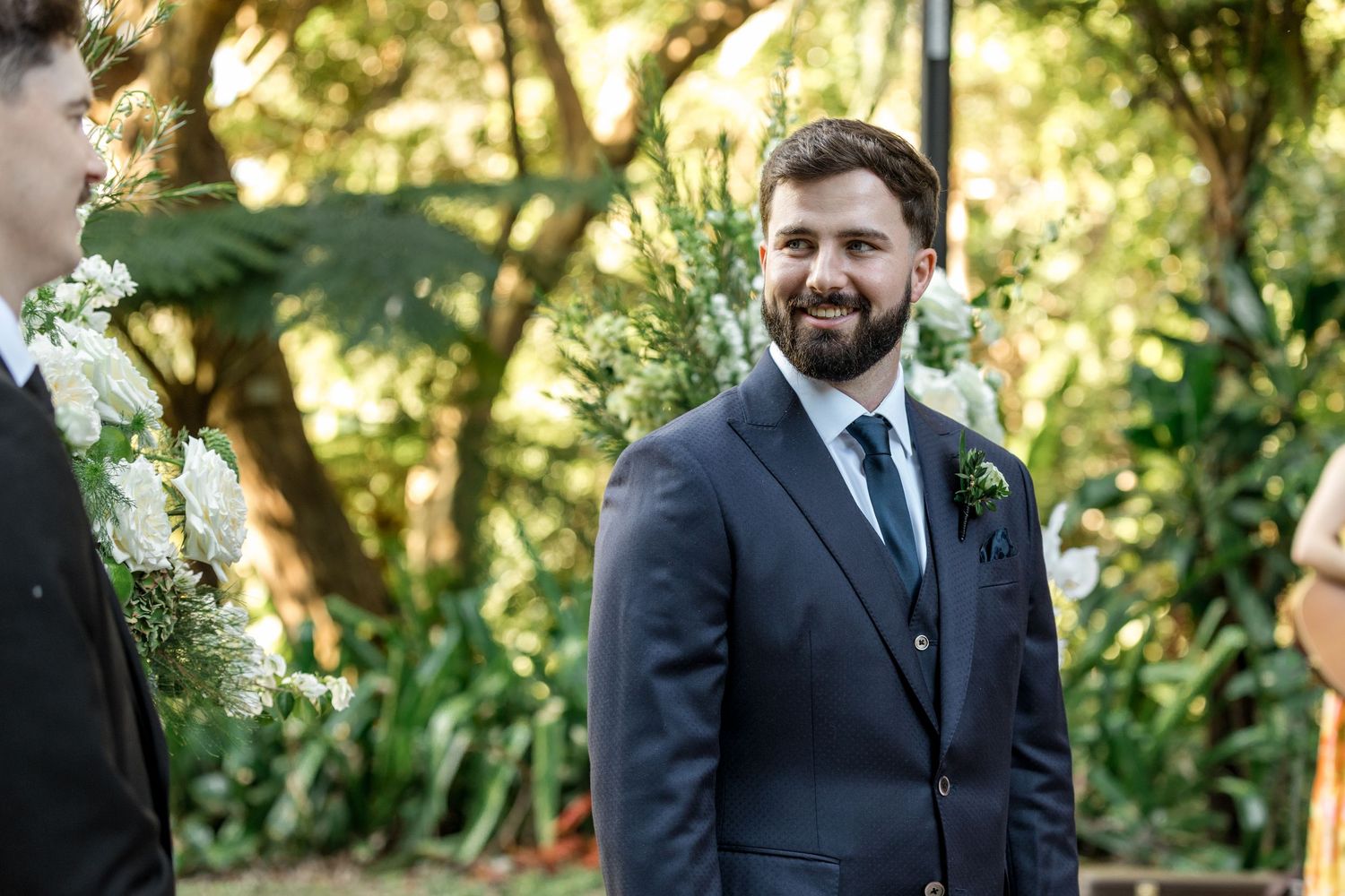 Smiling bearded groom in navy suit with boutonniere at outdoor garden wedding ceremony.