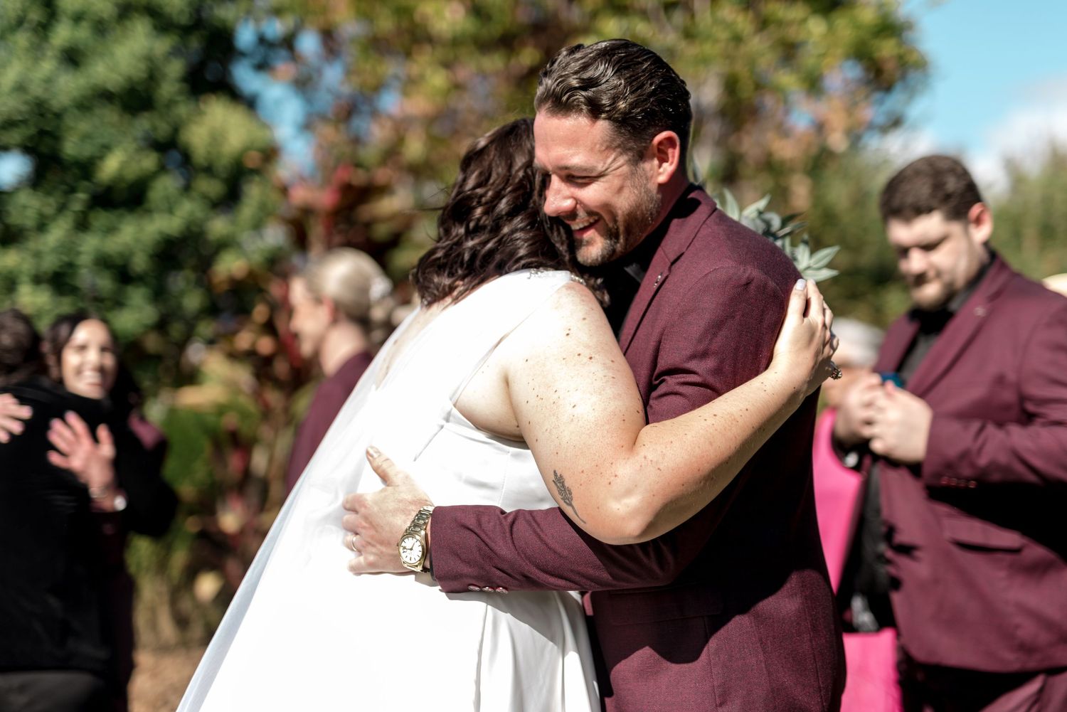 Groom in burgundy suit shares warm embrace with bride after outdoor wedding ceremony.