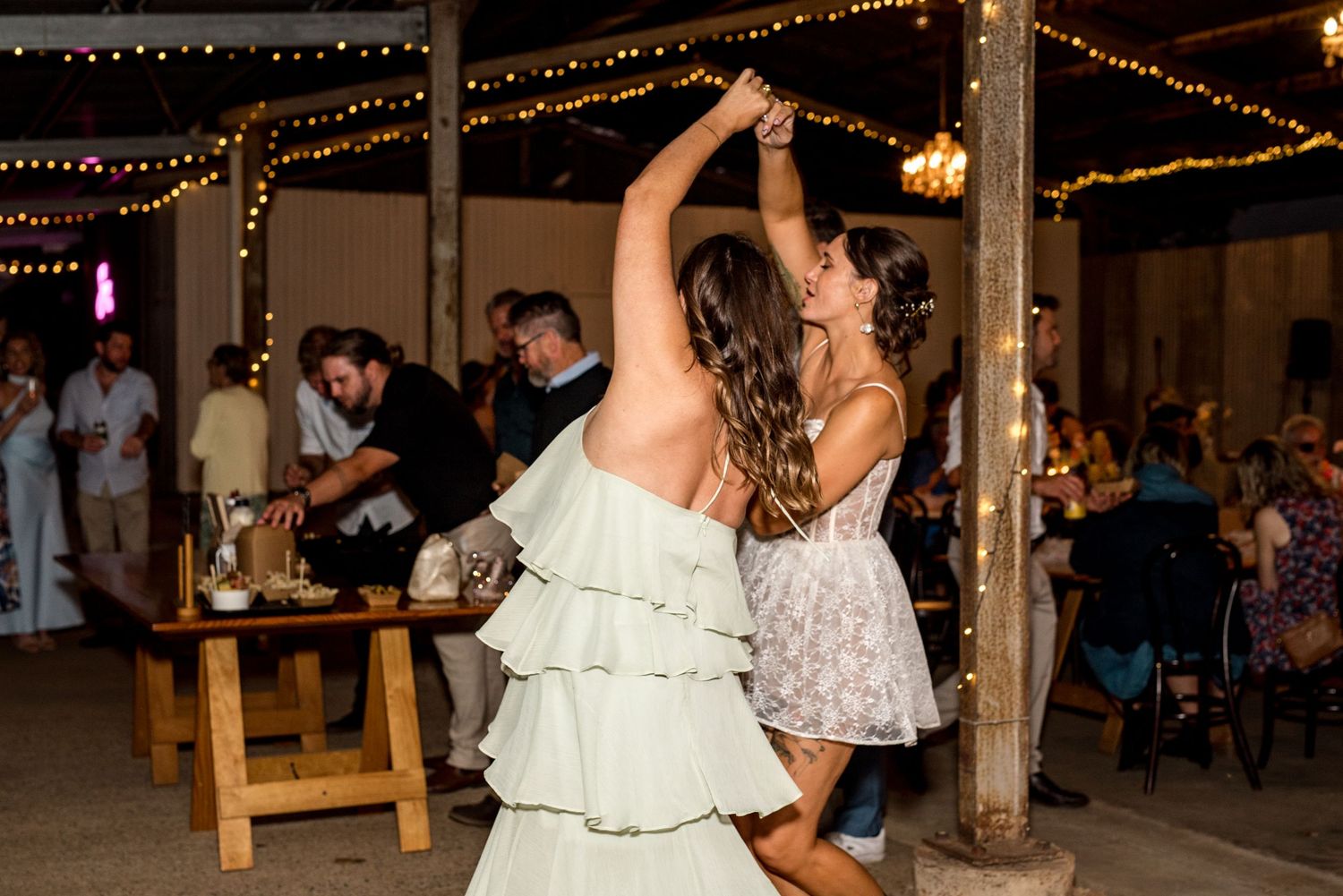 Two women dancing together at a rustic barn wedding reception with fairy lights overhead.
