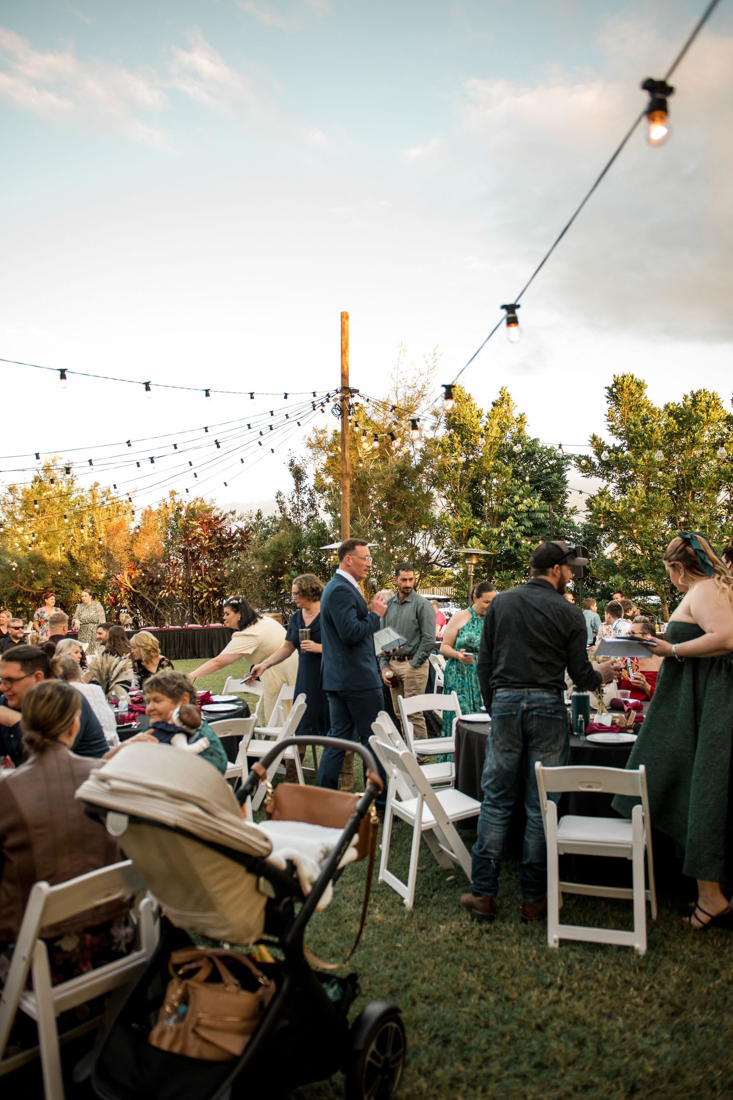 Outdoor wedding reception at dusk with string lights and guests mingling at tables.