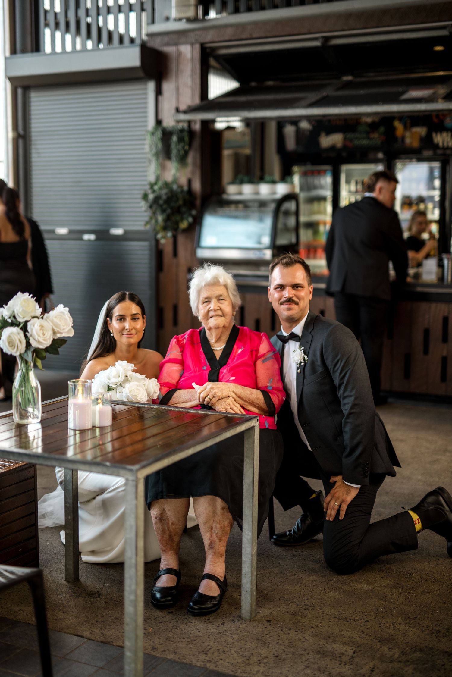 Bride and groom kneeling beside elderly grandmother at wedding reception table inside a warmly lit restaurant venue.