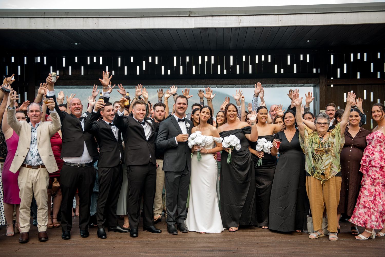 Joyful wedding party and guests celebrating with raised hands in a group photo at the reception.