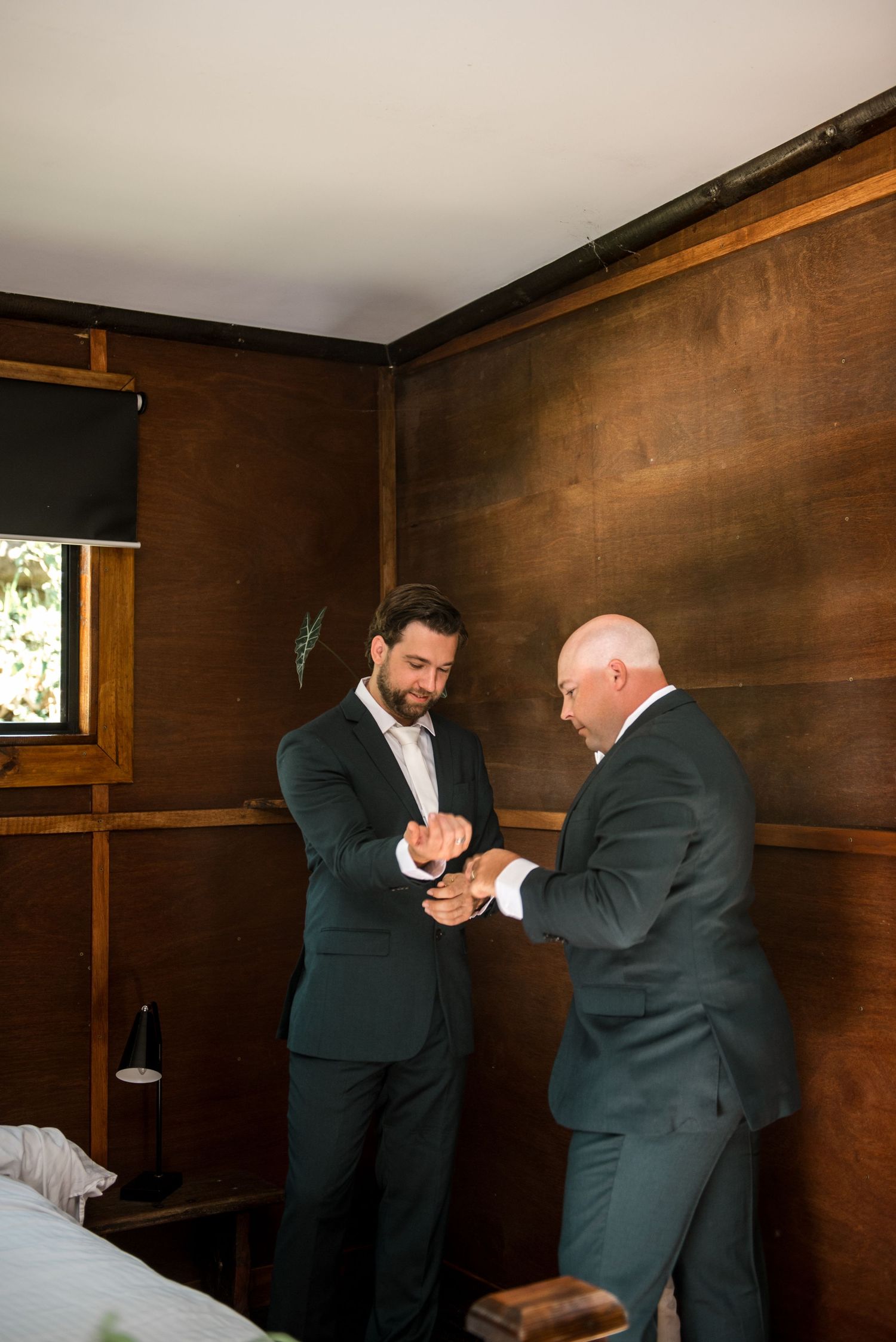 Two men in dark suits adjusting cufflinks together in a rustic wooden paneled room.