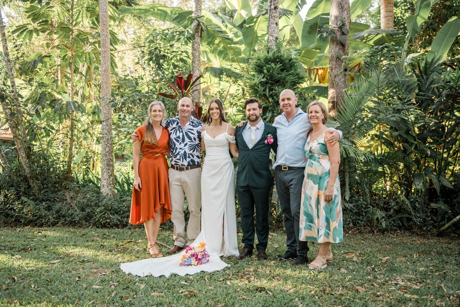 Newlyweds pose with six family members for group photo in lush tropical garden setting.