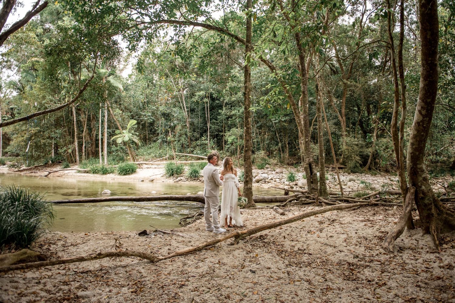 Bride and groom embrace near a peaceful forest stream surrounded by lush greenery.