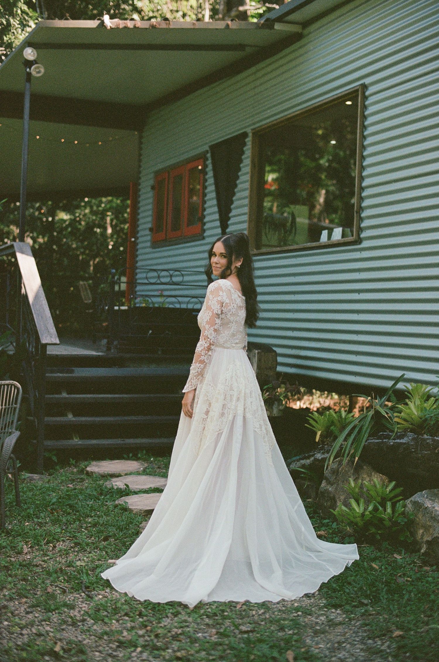 Bride in long-sleeve lace wedding gown posing outside rustic corrugated iron building.