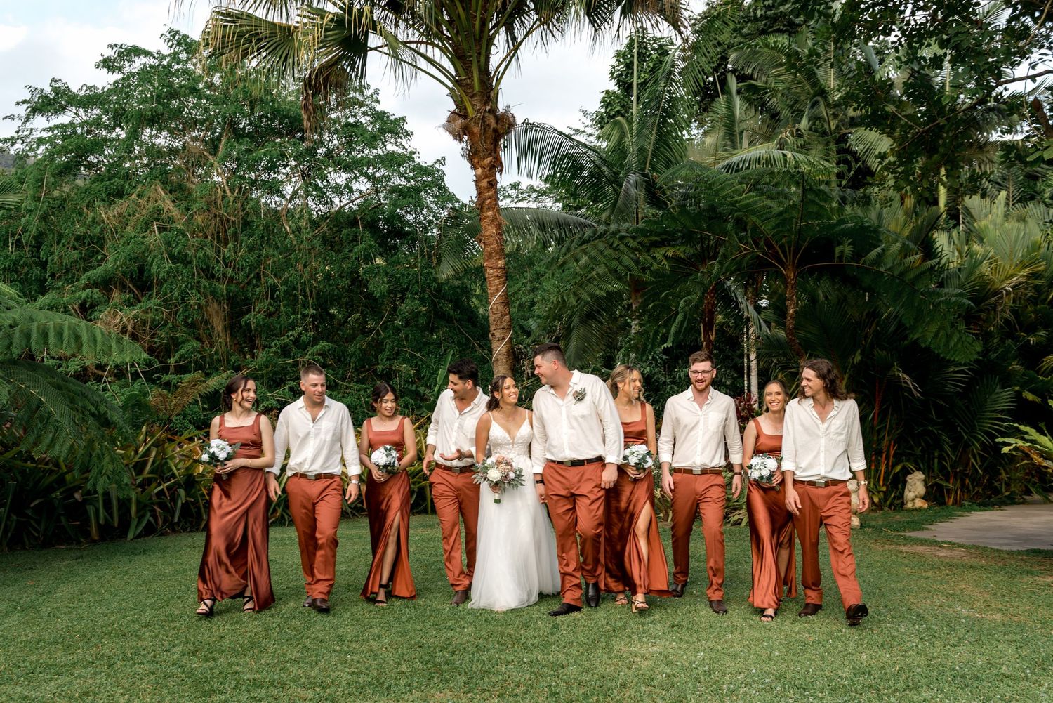 Wedding party walking together in lush tropical garden, bridesmaids in rust gowns and groomsmen in rust pants.