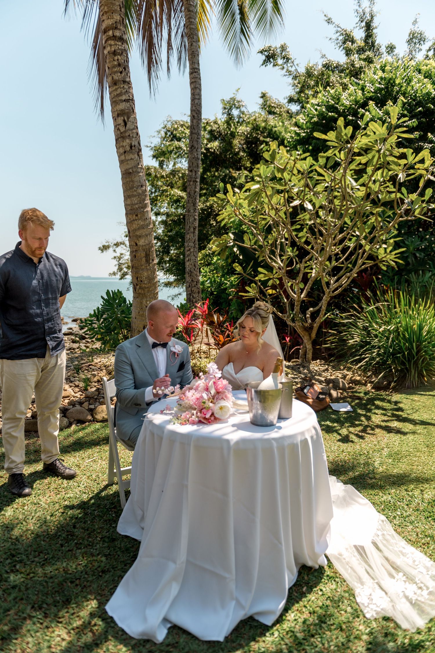 Bride and groom signing marriage certificate at white draped table in tropical garden setting by the sea.