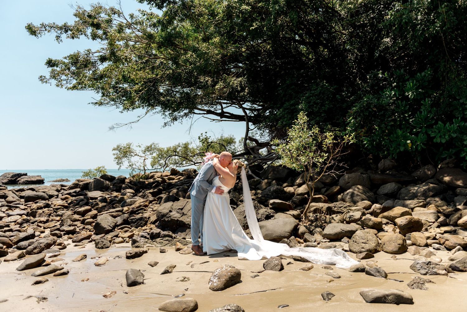 Wedding couple sharing a dip kiss on a rocky beach with lush tropical trees behind them.