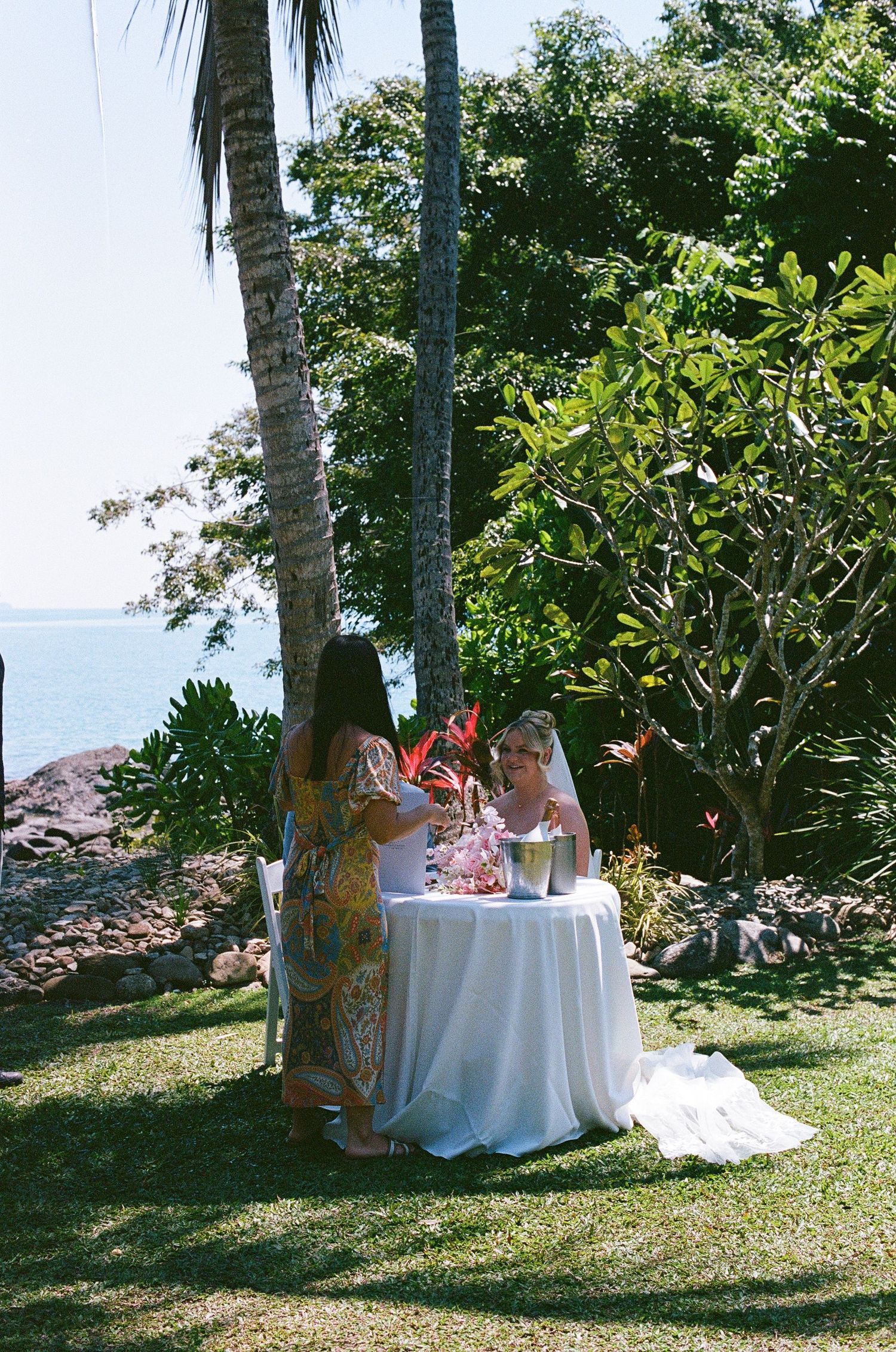 Bride in white gown sits at decorated table with guest in tropical garden near the ocean.