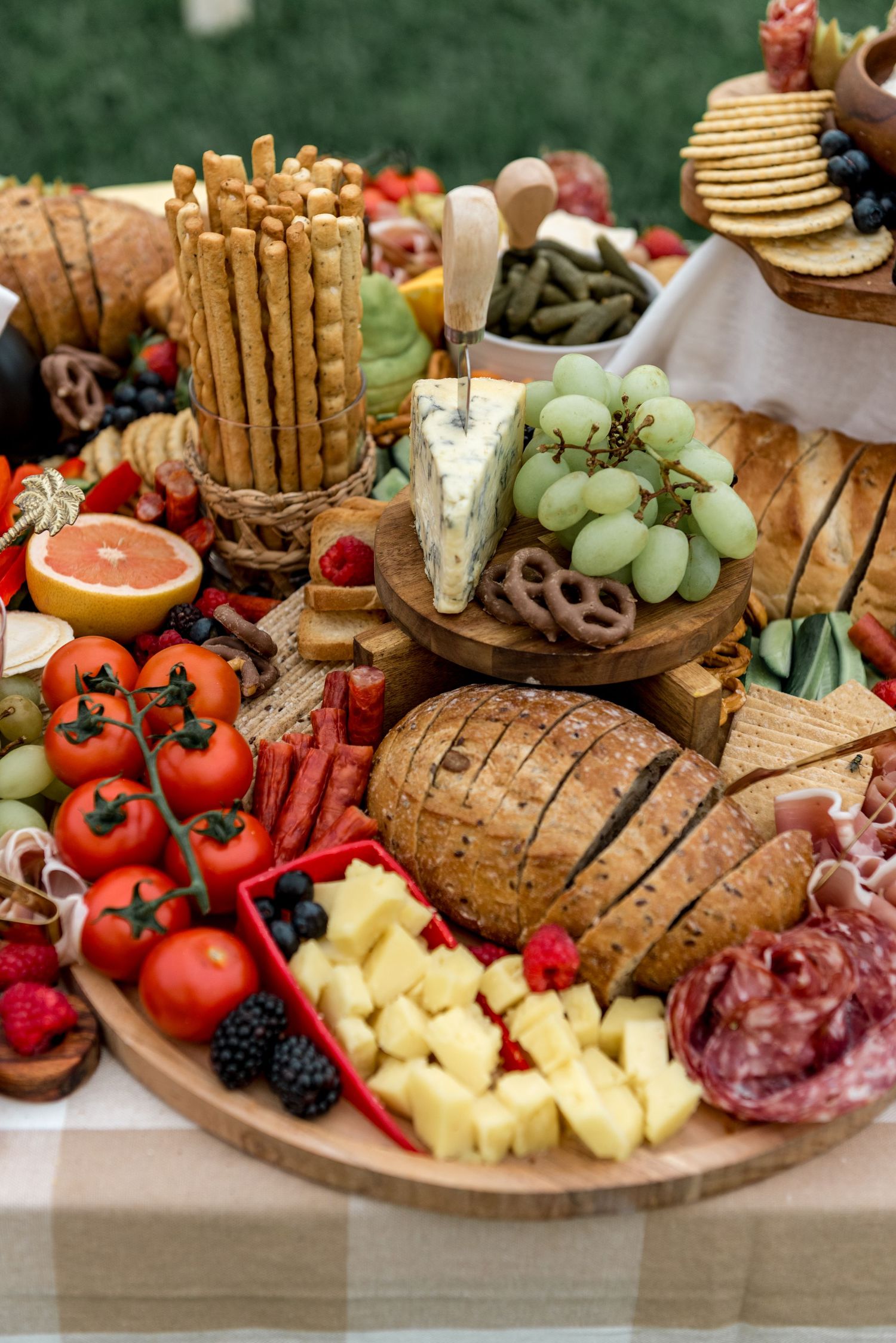 Rustic charcuterie board with cheeses, meats, grapes, tomatoes, breadsticks, and fresh fruits.