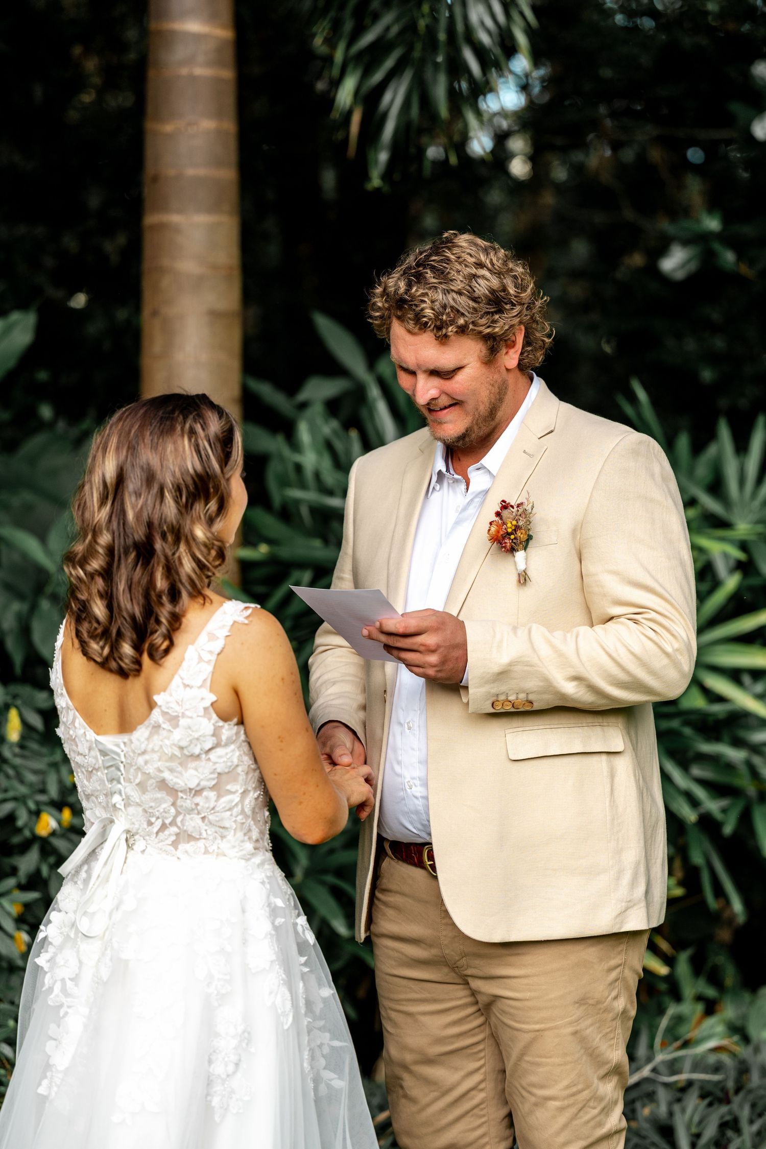 Groom reads wedding vows from card while holding brides hand in tropical garden ceremony.
