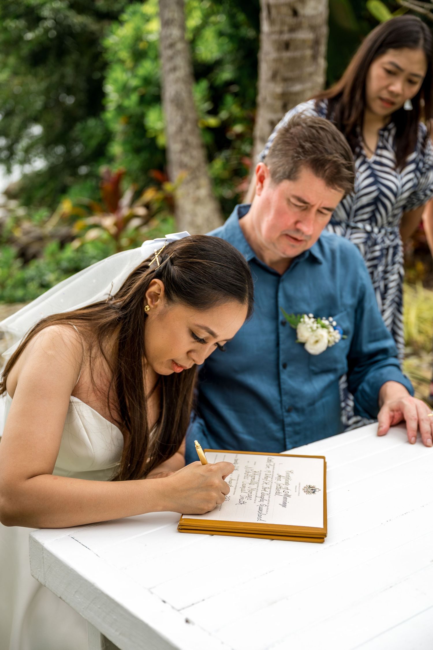 Bride signing wedding registry document at outdoor beach ceremony table with groom watching nearby at Little Cove, Port Douglas.