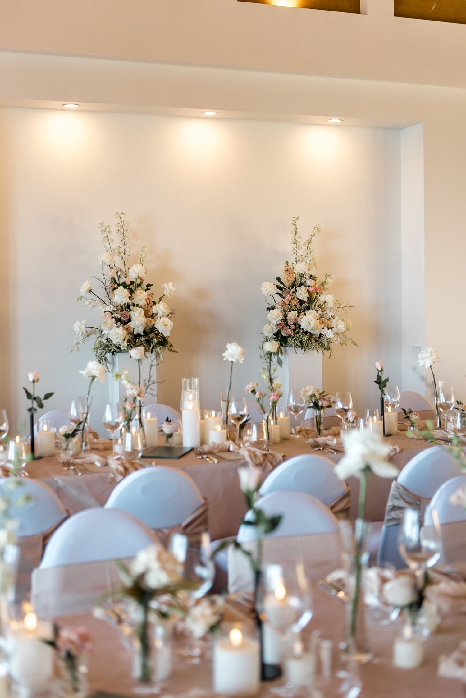 Wedding reception hall with tall floral arrangements, white chair covers, candles, and single stem roses on long tables.