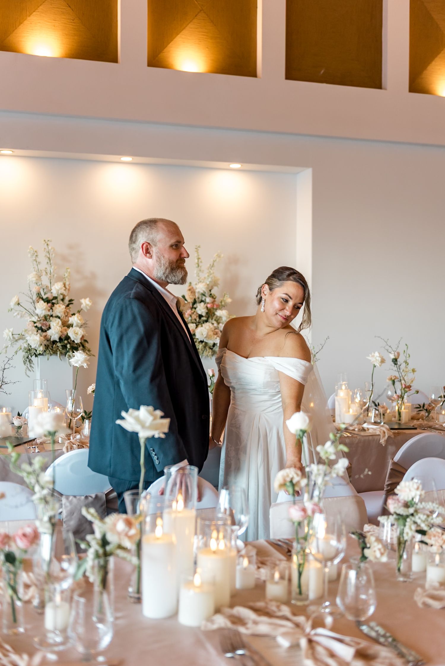 Bride laughs joyfully while holding groom's hand at their candlelit wedding reception with floral arrangements.
