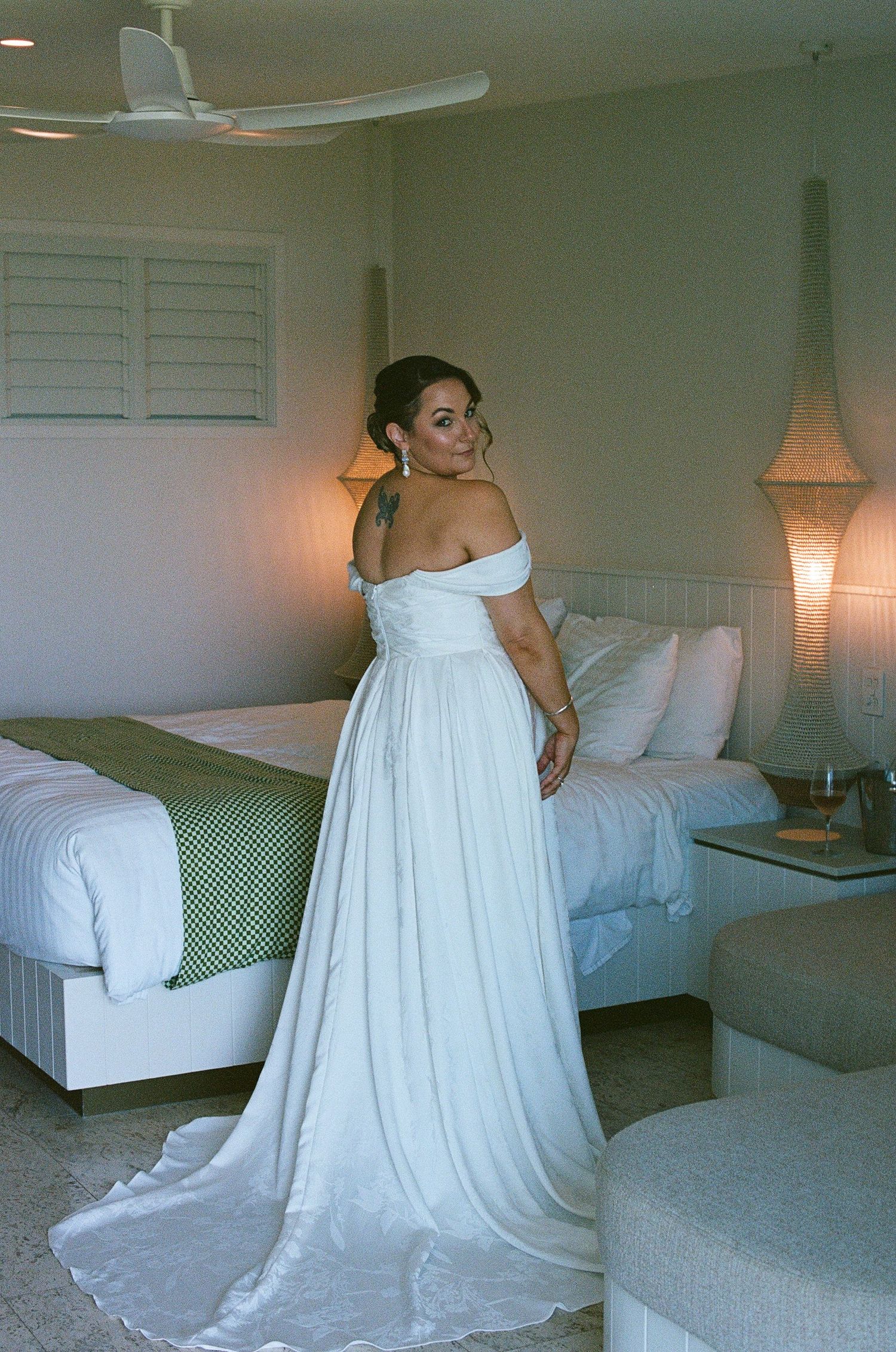 Bride in elegant white off-shoulder wedding gown with train posing in a tropical resort hotel room.