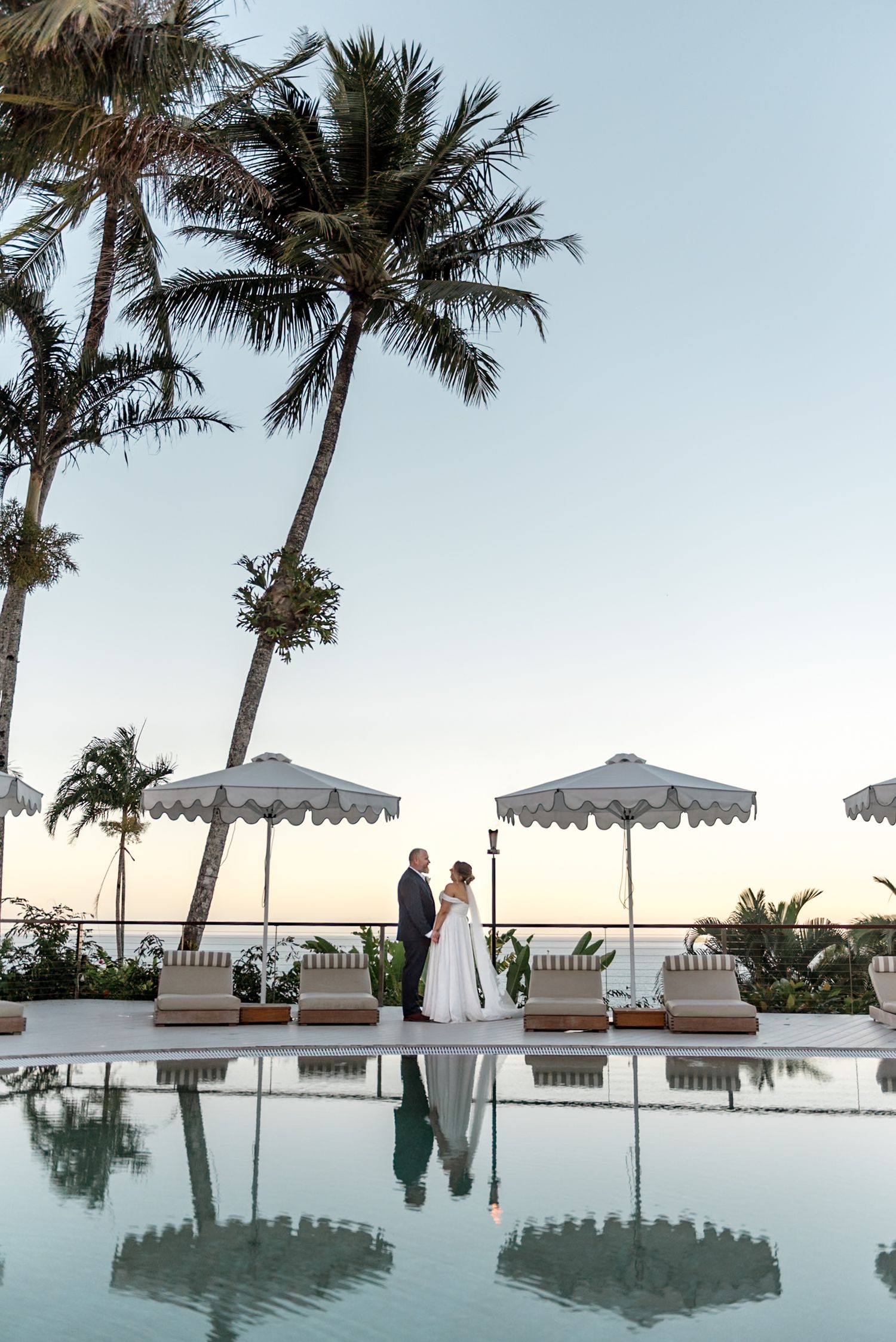 Wedding couple stands by a tropical resort pool at dusk with palm trees and ocean horizon backdrop.