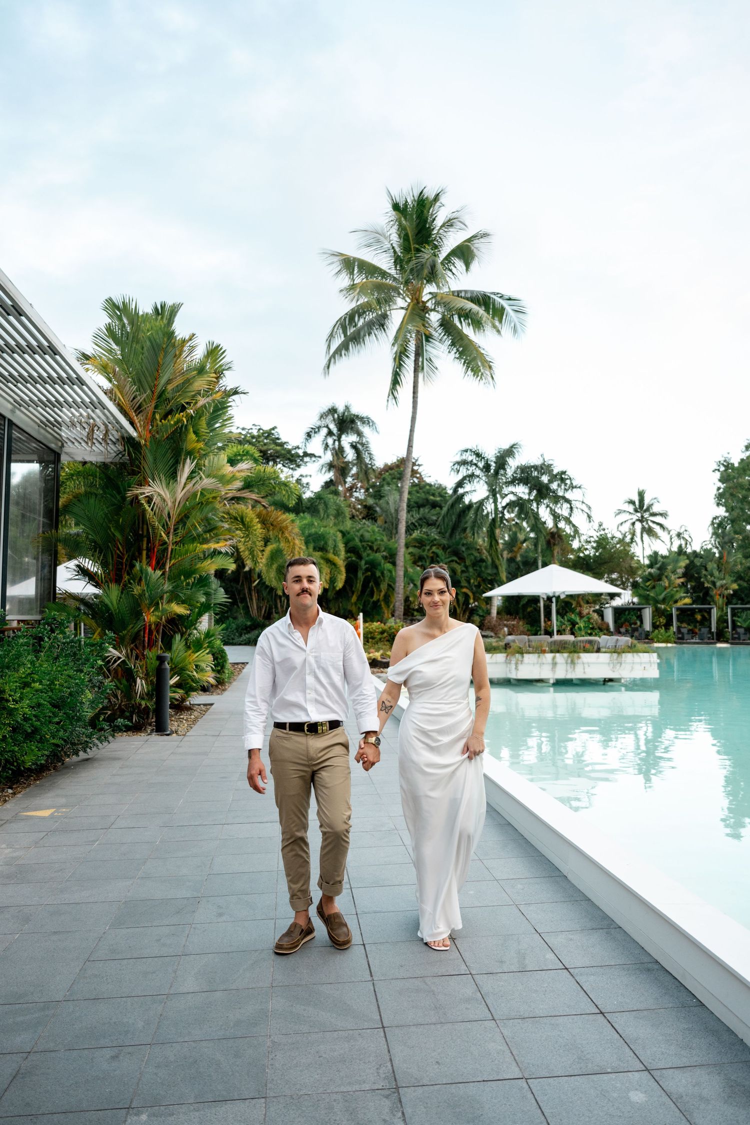 Smiling bride and groom walking hand in hand along a tropical resort pool in white attire at Sheraton Grand Mirage Port Douglas.