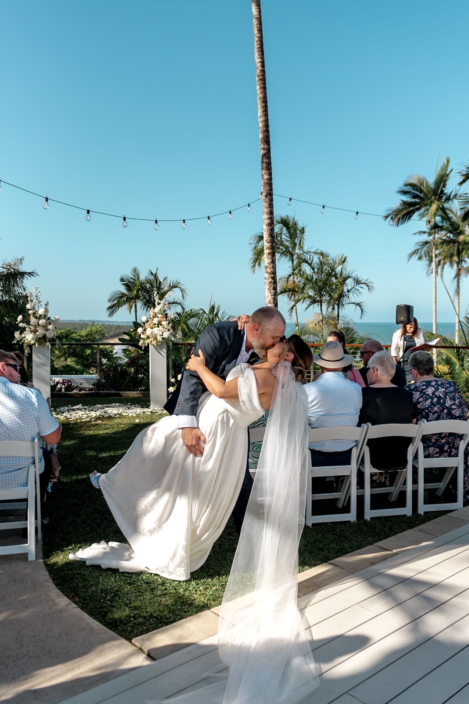 Groom lifts and kisses bride at tropical outdoor wedding ceremony with palm trees and ocean backdrop.