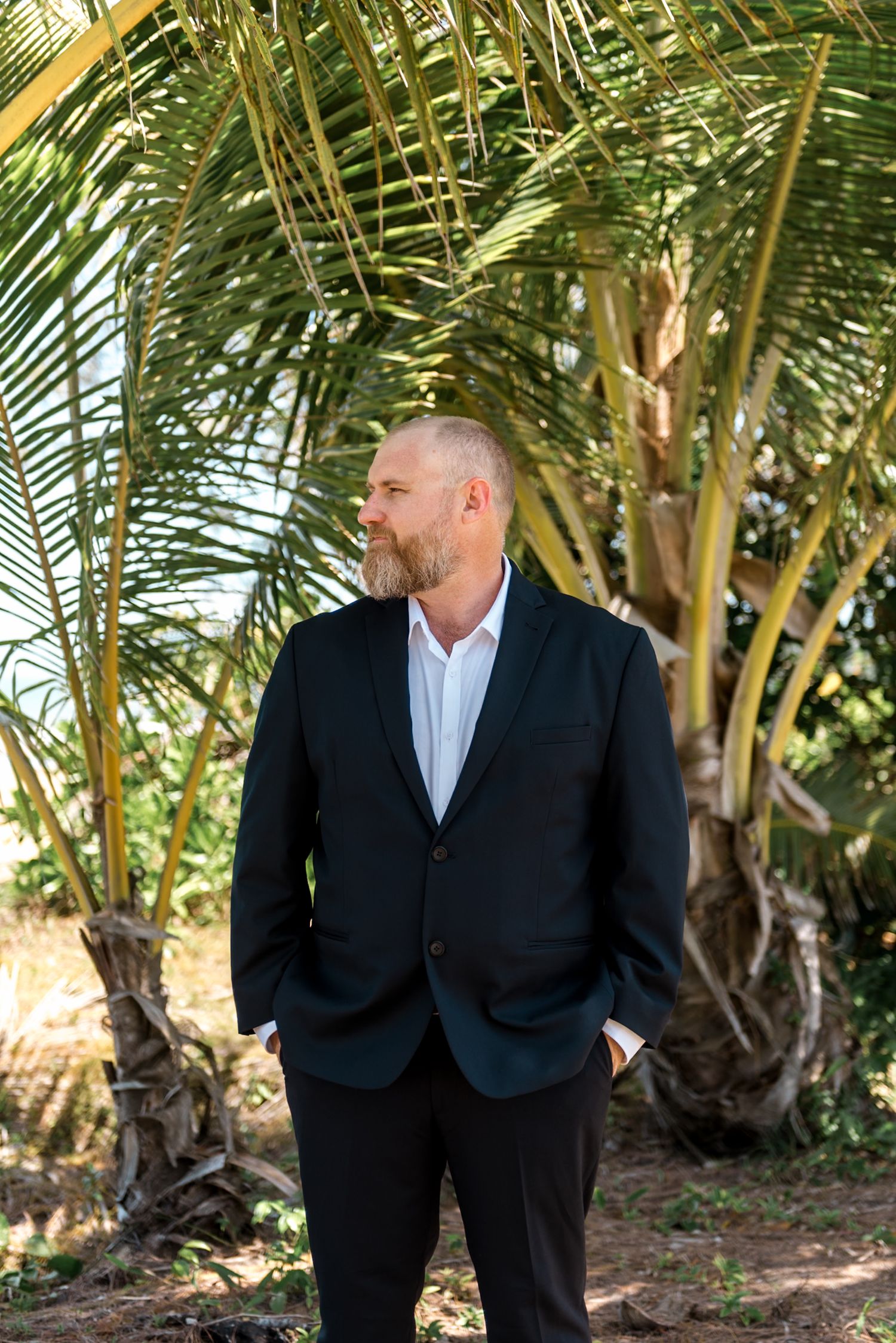 Bearded man in a dark blazer standing outdoors beneath lush green palm tree fronds.