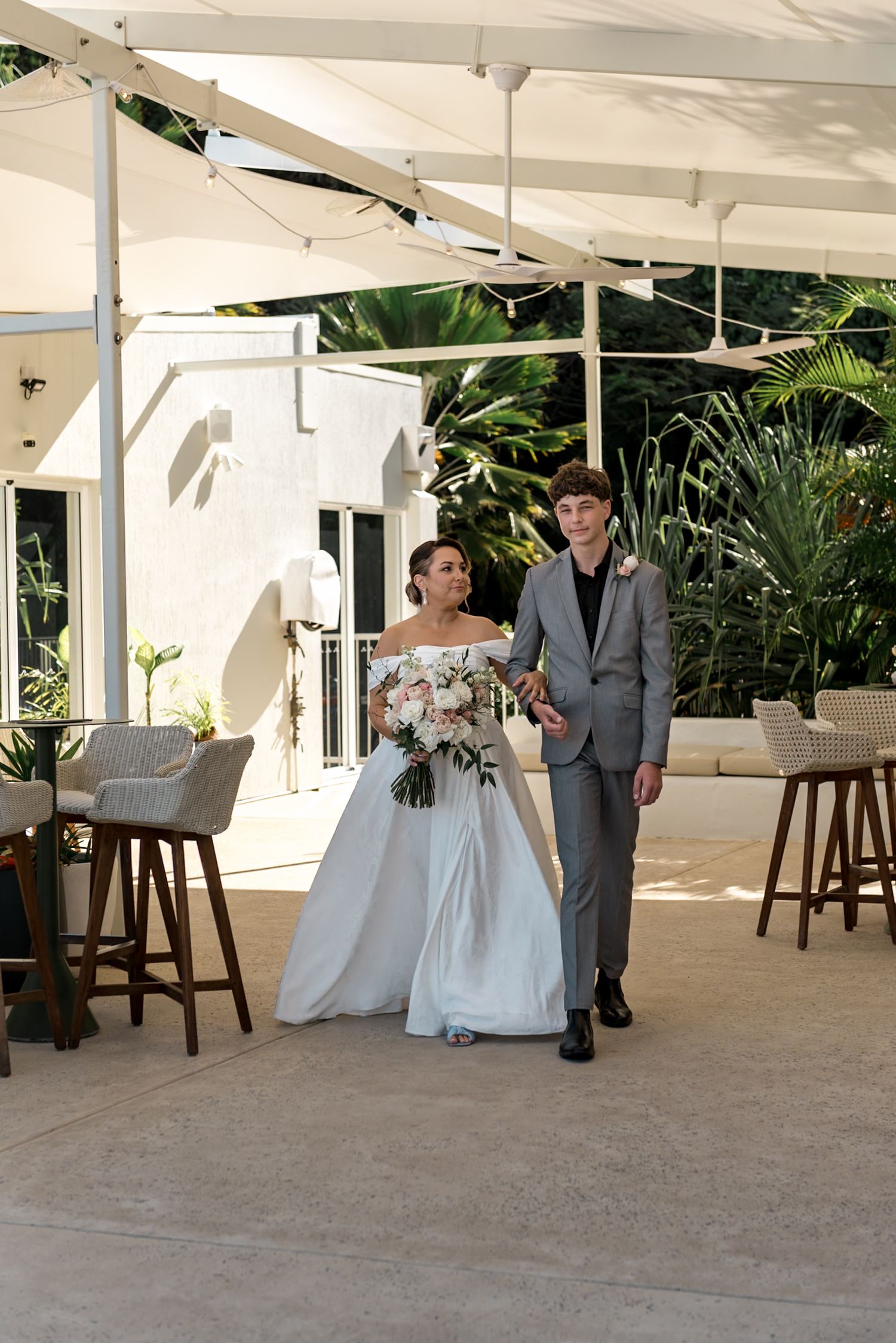 Bride in off-shoulder white gown walks with groom in gray suit at outdoor wedding venue with tropical greenery.