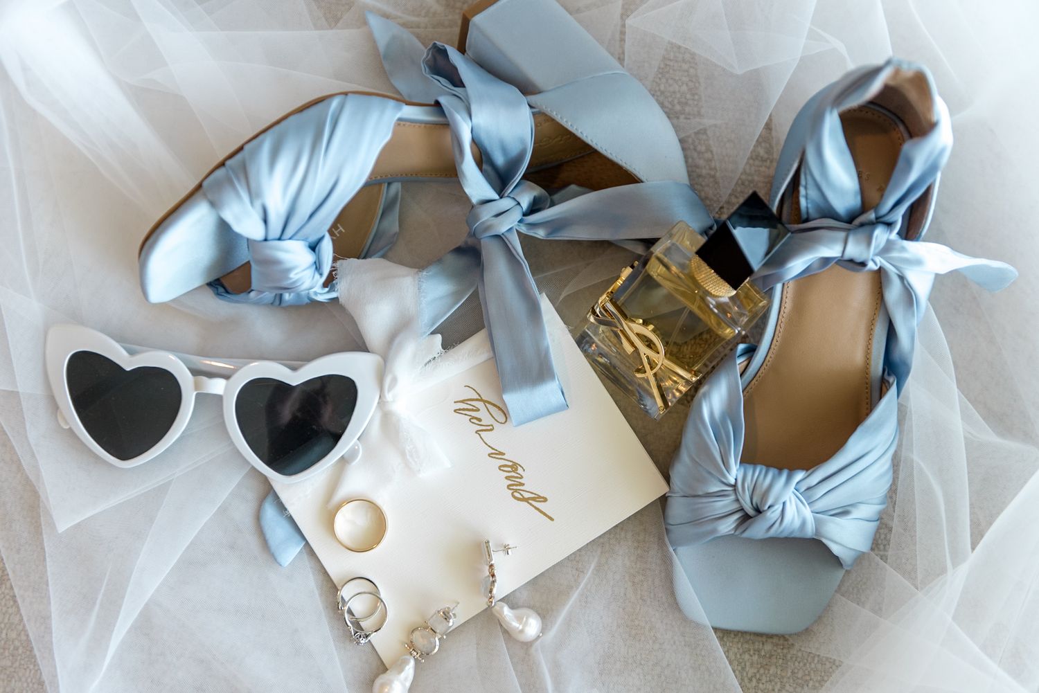 Bridal flat lay featuring blue satin heels, heart sunglasses, pearl earrings, and YSL perfume on tulle.
