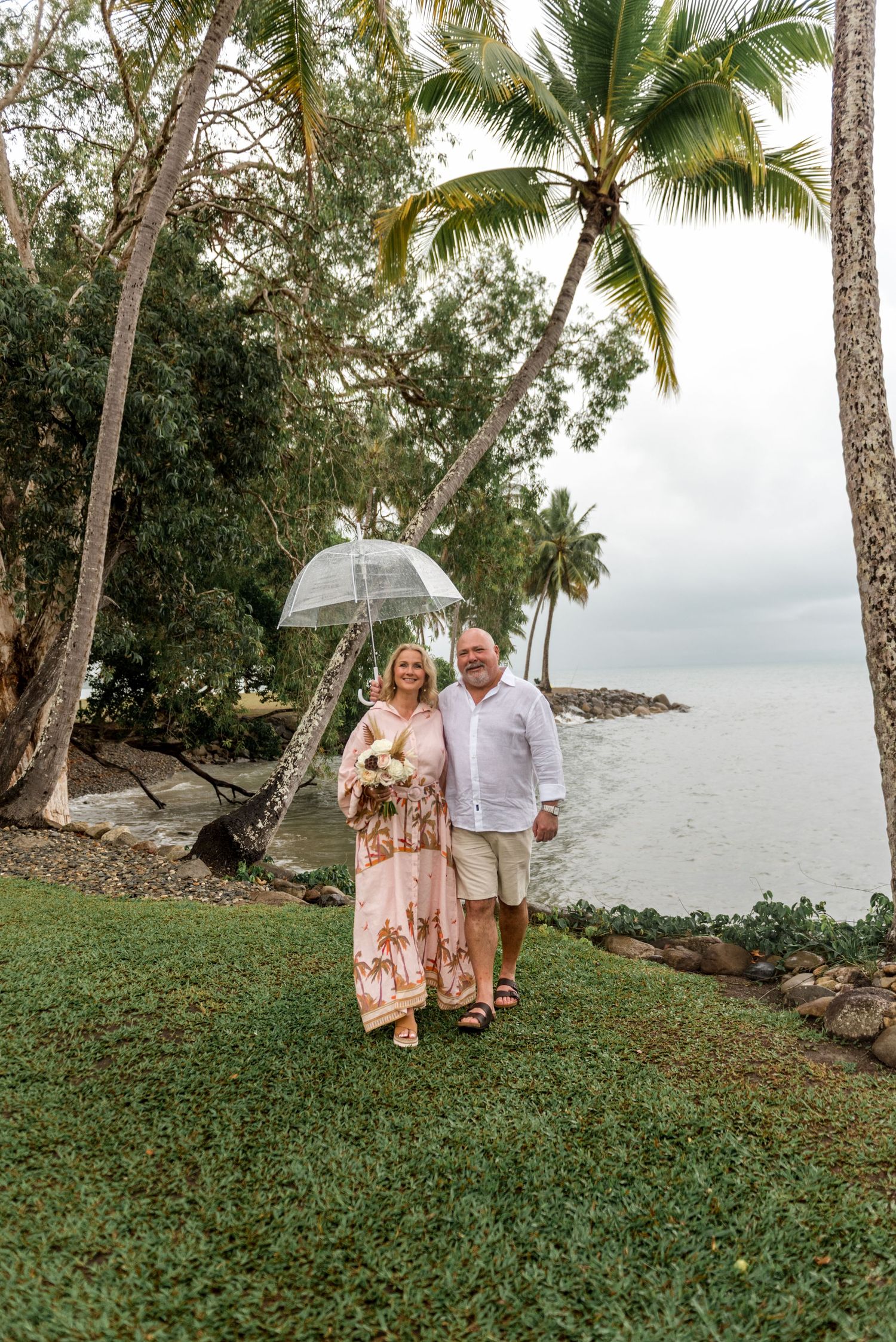 Couple walking hand in hand under a clear umbrella by a tropical palm-lined waterfront on a cloudy day.