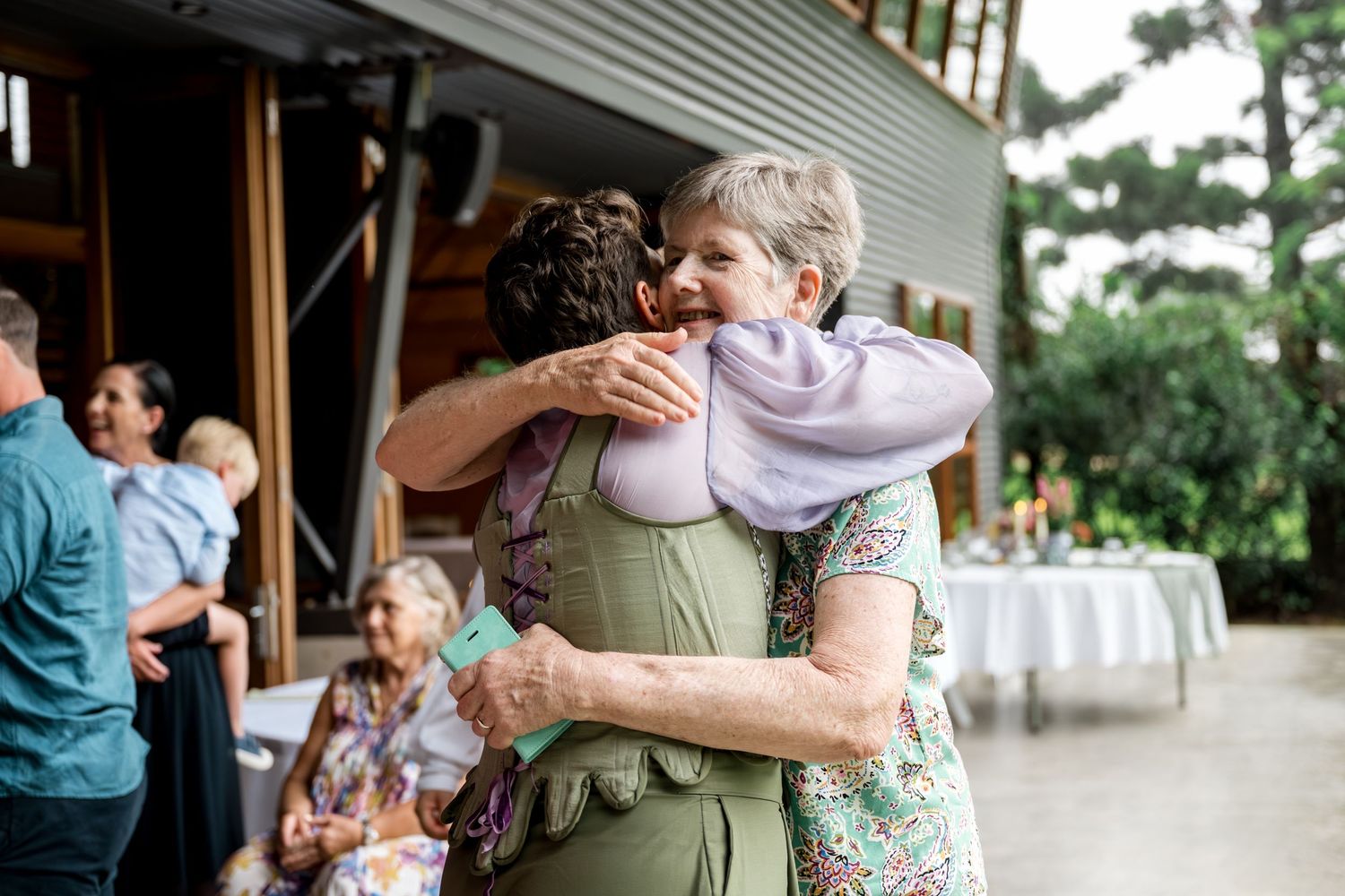 Older woman and guest share a joyful hug at an outdoor wedding celebration.