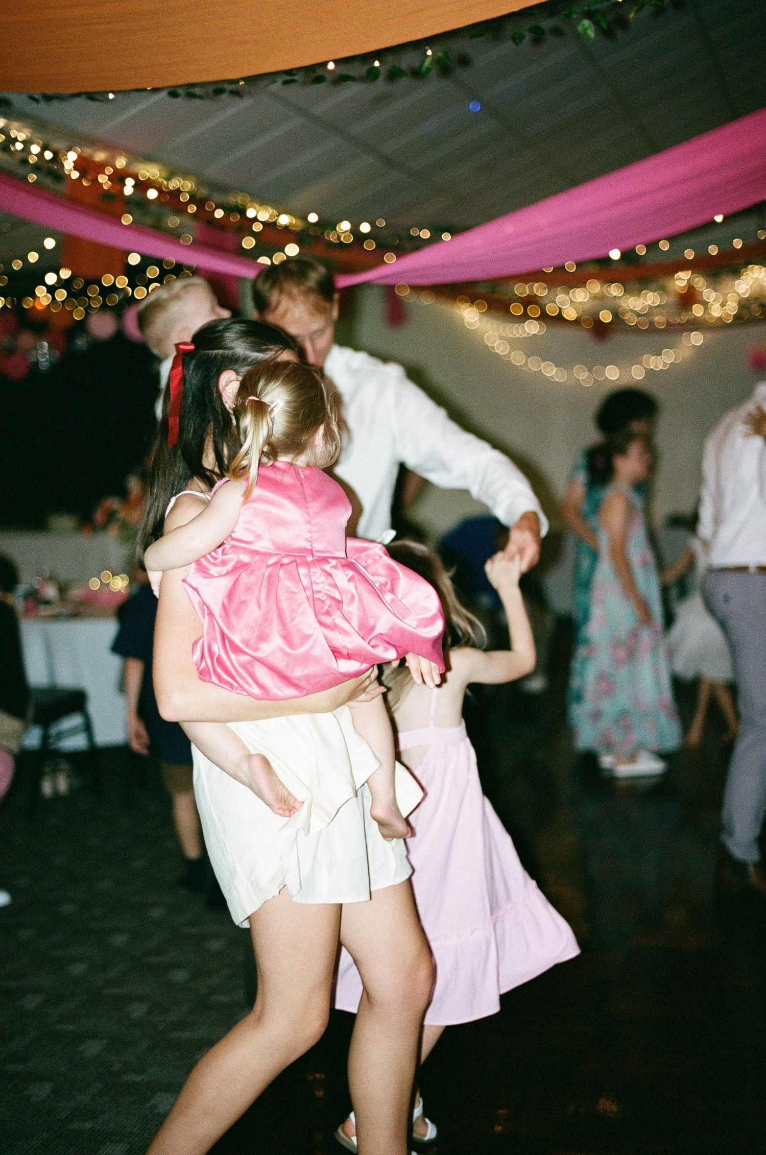 Children and adults dancing together under pink drapes and string lights at a wedding reception.