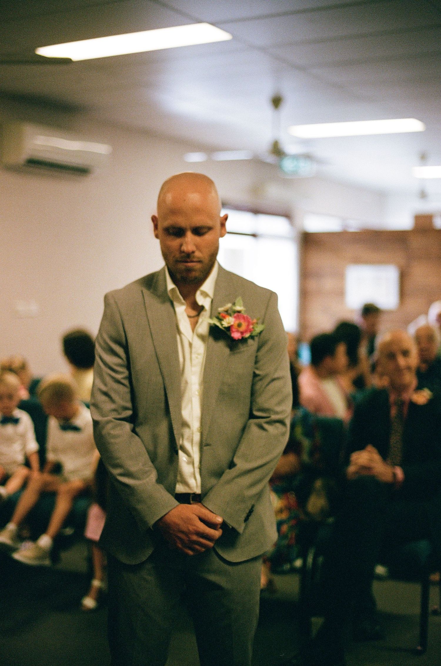 Bald groom in gray suit with pink boutonniere stands solemnly during indoor wedding ceremony awaiting his bride.