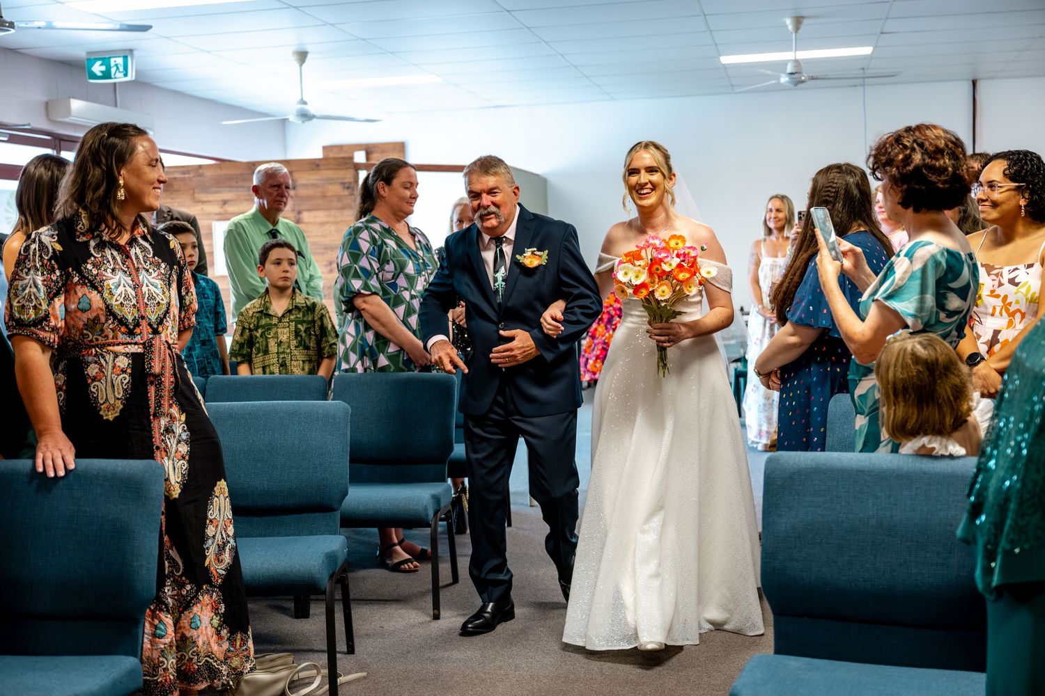 Bride in white gown walks down aisle with father while guests celebrate at indoor wedding.