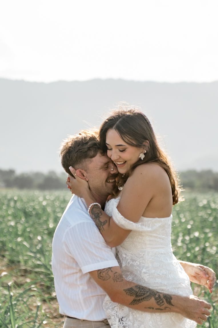 Laughing bride being held by tattooed groom in a sunny green field wedding portrait session.