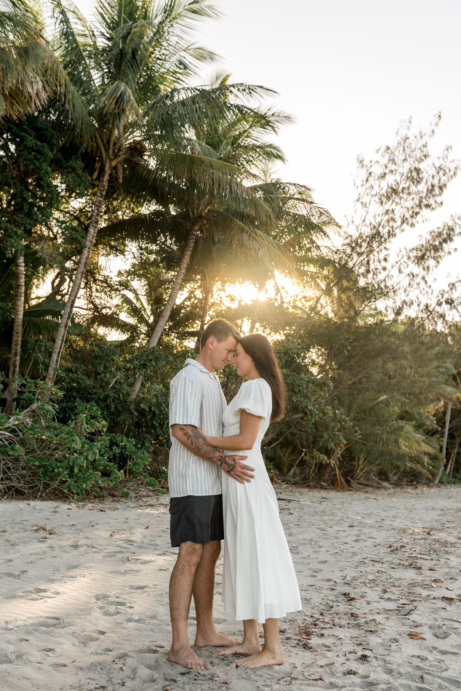 Romantic couple touching foreheads at sunset on tropical beach surrounded by golden-lit palm trees.