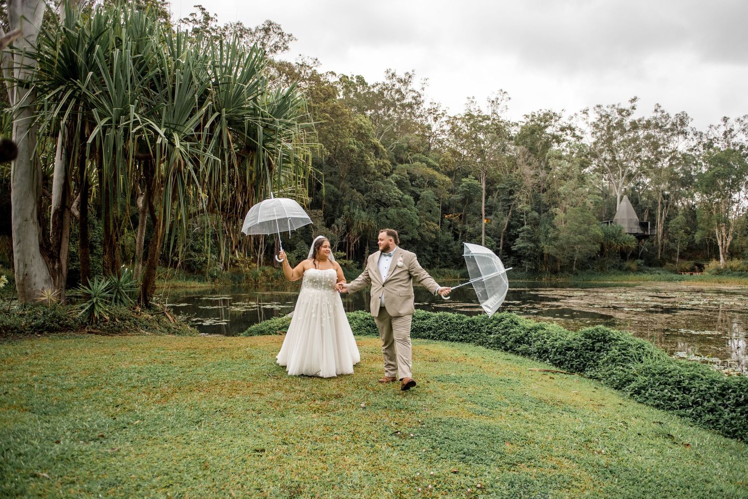 Joyful bride and groom walk hand in hand holding clear umbrellas through a lush green garden.
